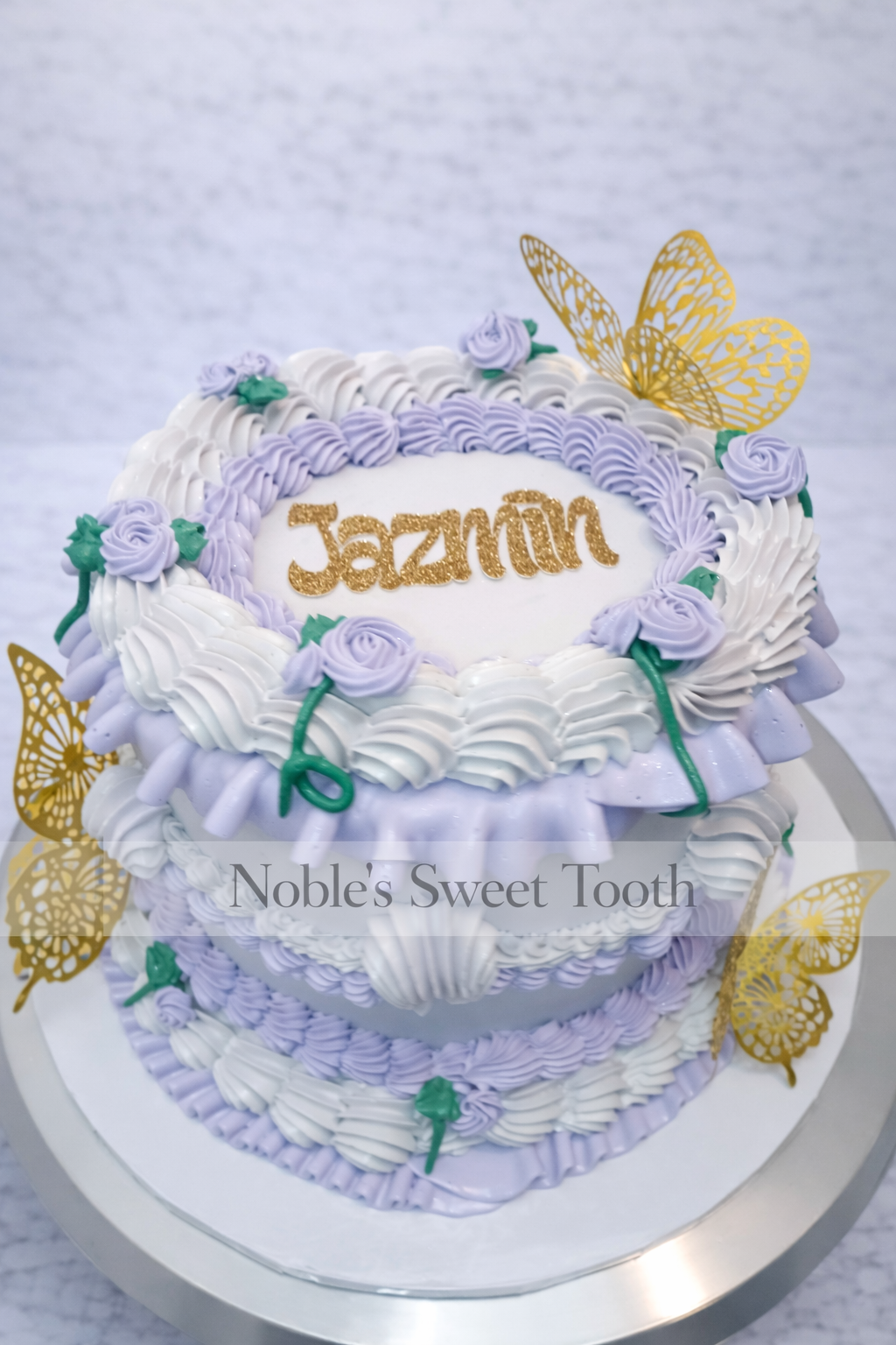 A two-tiered cake decorated with white and lavender frosting, purple roses, green leaves, and gold butterfly toppers. The top of the cake has the name "Jazmin" written in gold glitter icing.