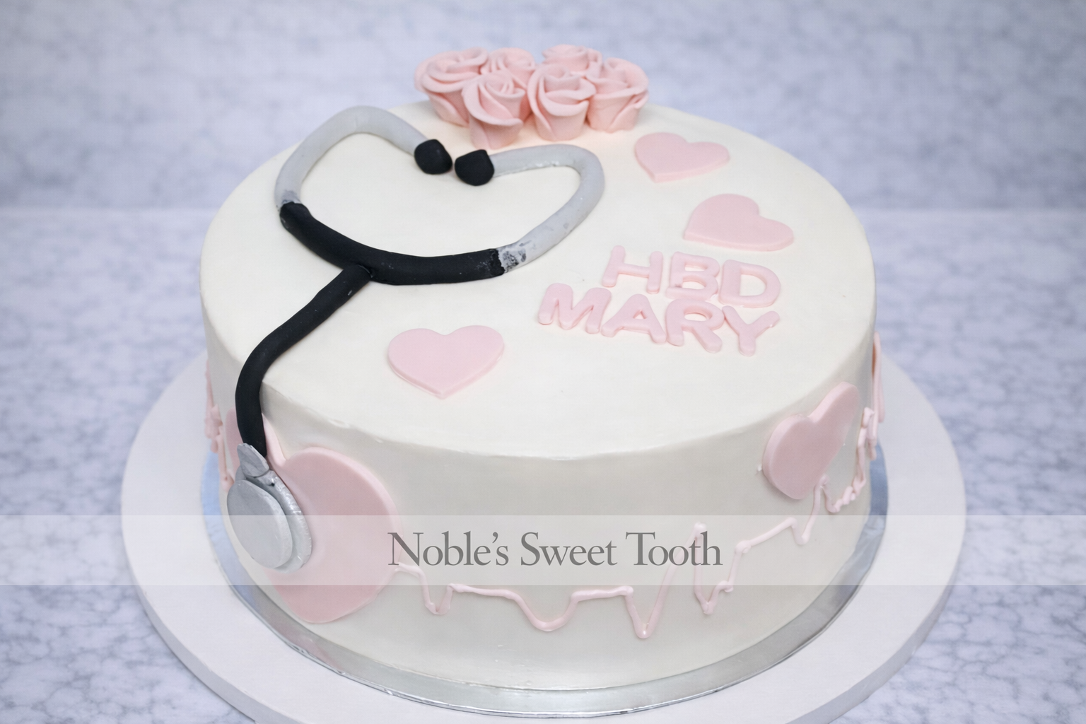 A decorated birthday cake with a stethoscope and pink hearts, inscribed with 'HBD MARY', on a white cake board.