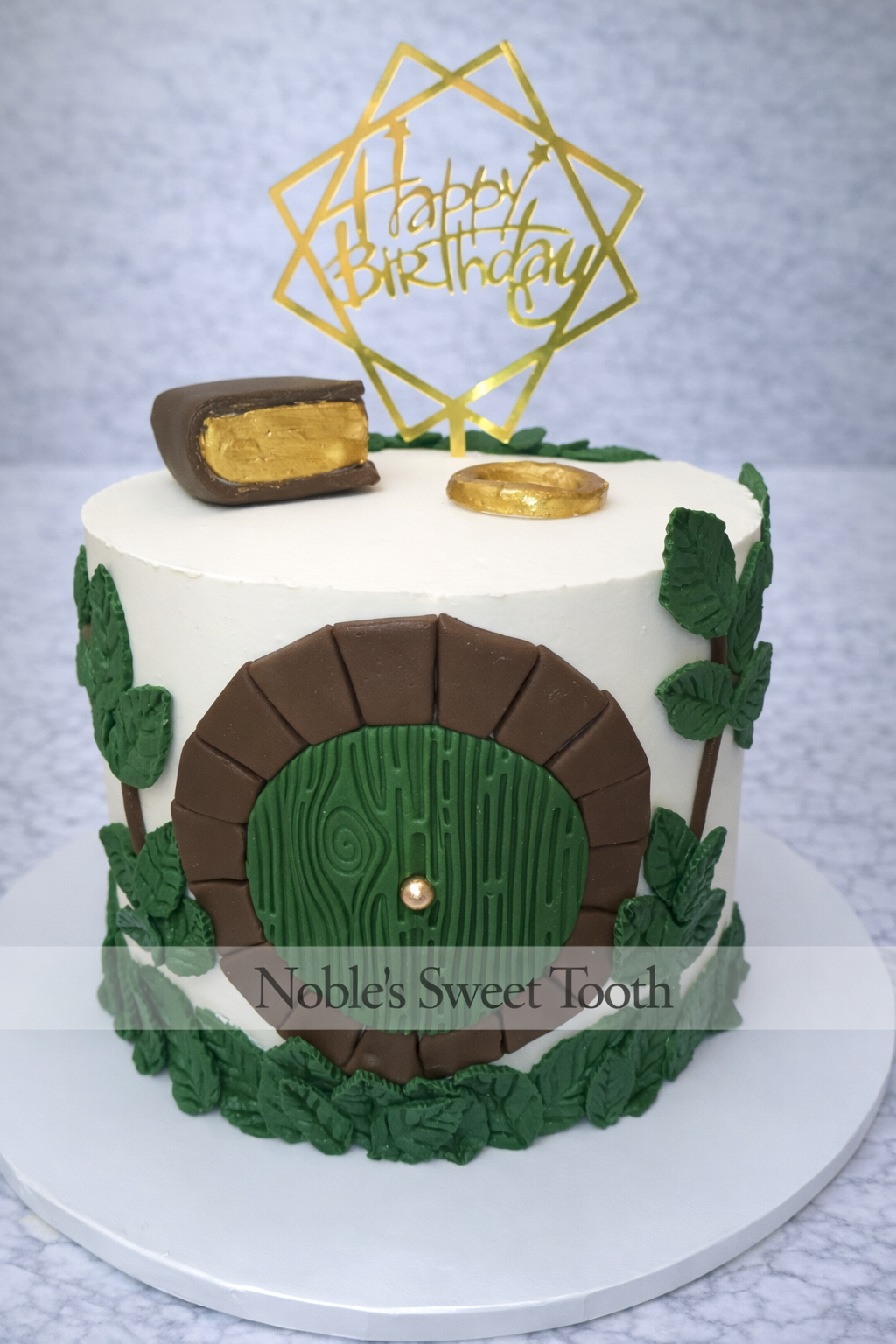 A cake decorated like a house with a green door, surrounded by green leaves, topped with a 'Happy Birthday' topper, and featuring a chocolate and gold coin on top. The cake is labeled 'Noble's Sweet Tooth'.