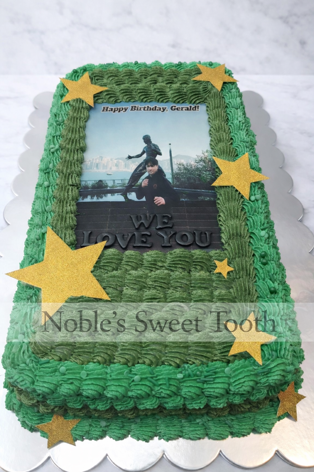 A rectangular birthday cake with green frosting and gold star decorations, featuring a printed photo of a young person sitting on stairs by a statue with city and water in the background. The cake has a message that says, 'WE LOVE YOU,' and a small t
