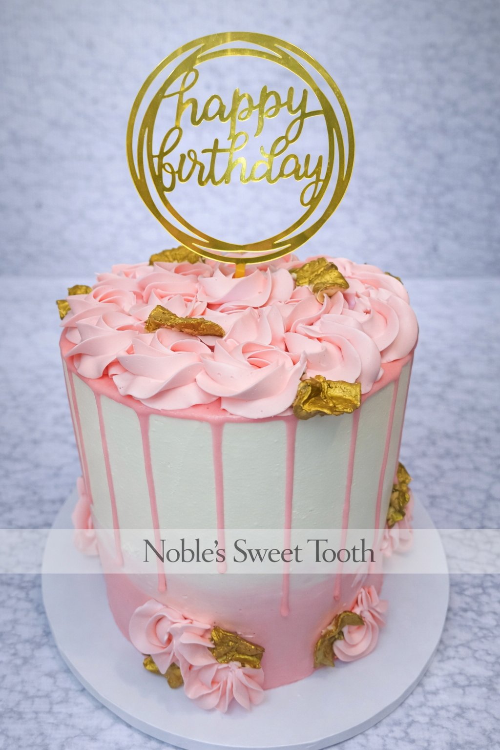 A tall birthday cake decorated with pink frosting and gold accents, topped with a gold 'happy birthday' cake topper, on a white round cake board.