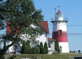 A lighthouse with a red and white exterior near a house with a red roof, surrounded by greenery under a clear sky.