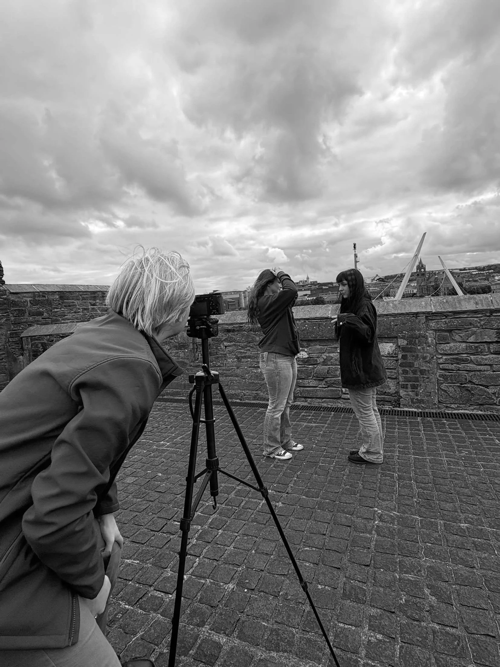 Two women standing on a rooftop, one adjusting her hair and the other speaking, while a person with light-colored hair and a jacket is observing and recording them with a camera on a tripod under an cloudy sky.