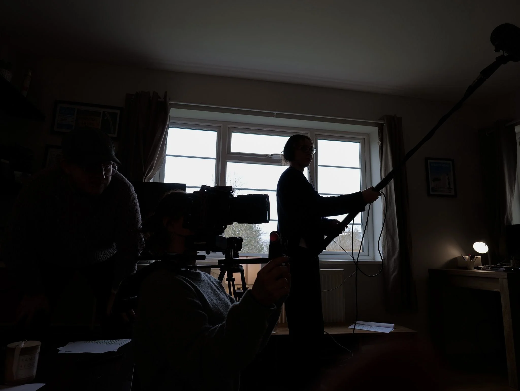 Silhouette of a person holding a boom microphone and a person sitting at a camera setup in a room with windows, curtains, and a desk with a lamp.