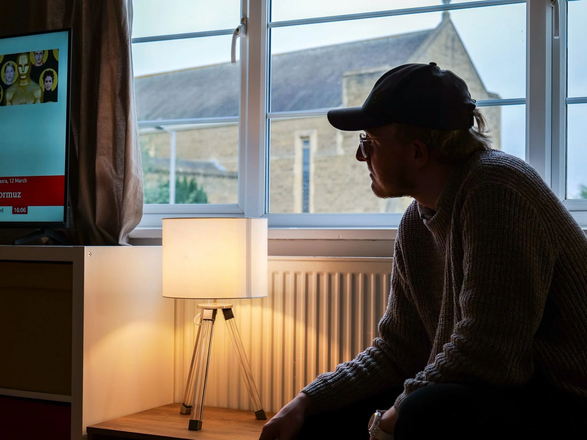 A man sitting indoors next to a window and a lamp, watching a television screen.