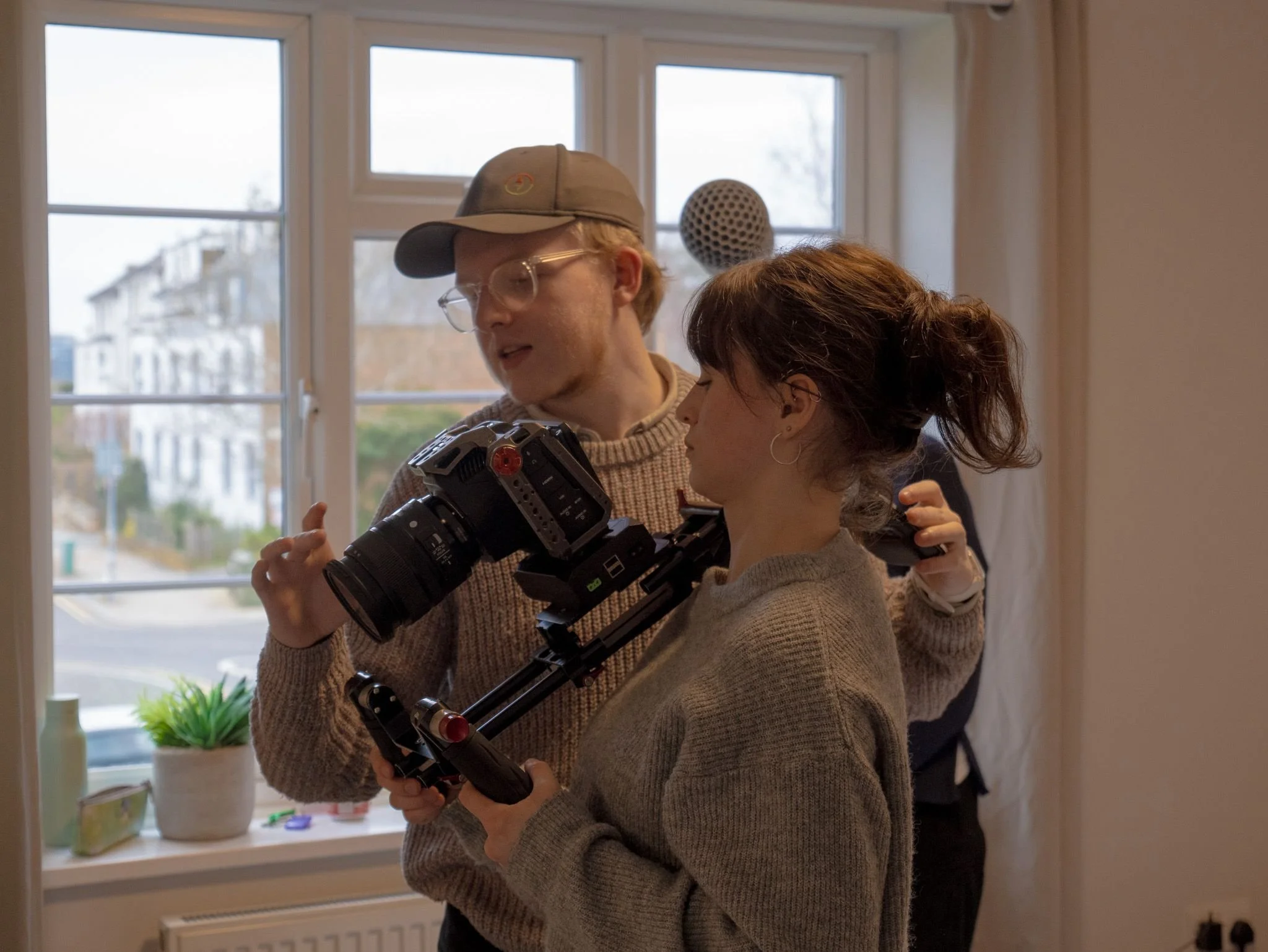 Two people, a man with glasses and a woman, working with a camera in a room with big windows and a view of a building outside.