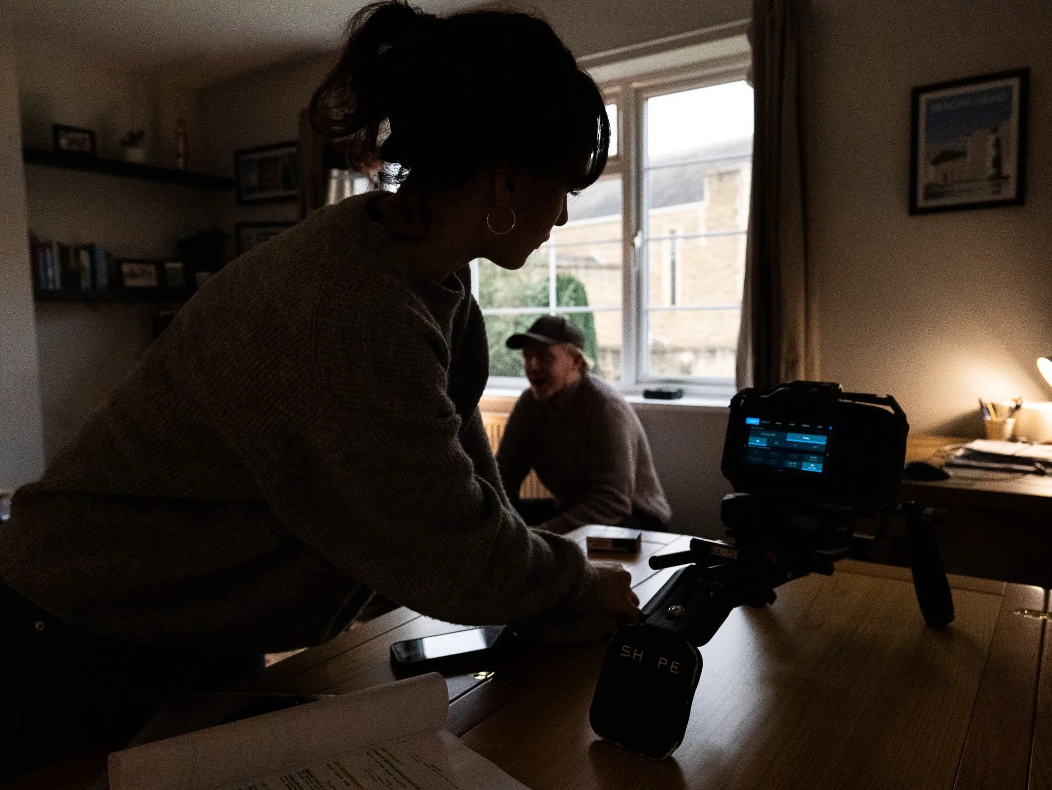 Two people in a dimly lit room, one handling a camera on a stabilizer, with a window behind them and another person sitting at the table with a hat, smiling.