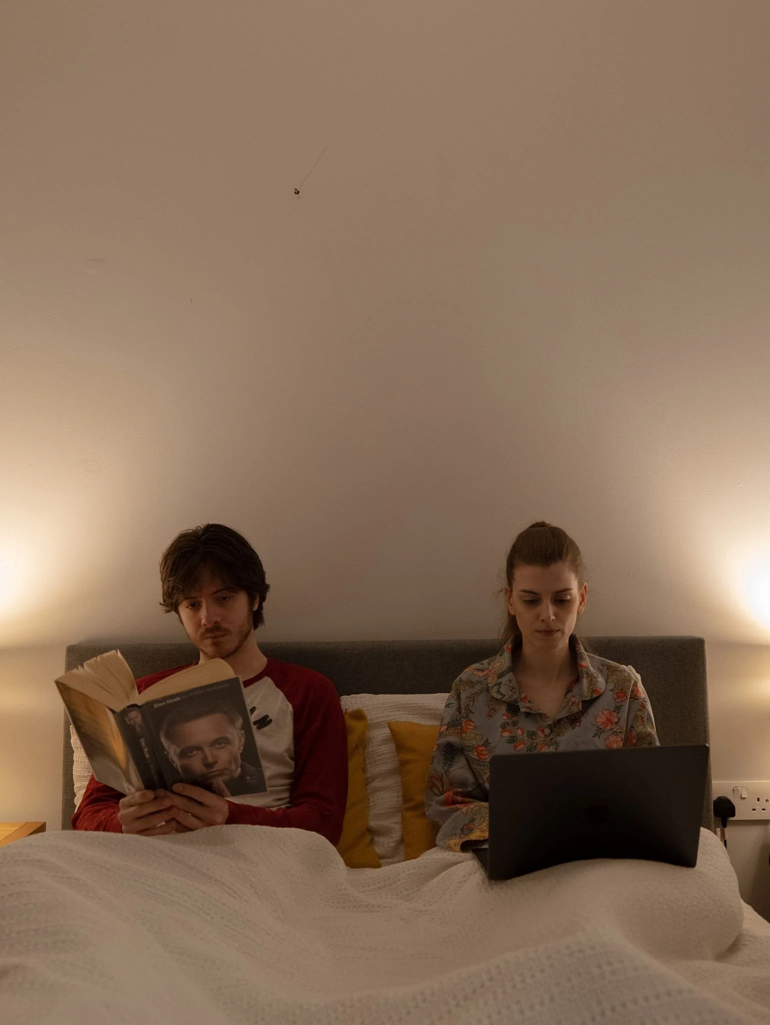 A man and a woman sitting in bed with a white blanket, each engaged in different activities; the man is reading a book, the woman is using a laptop.