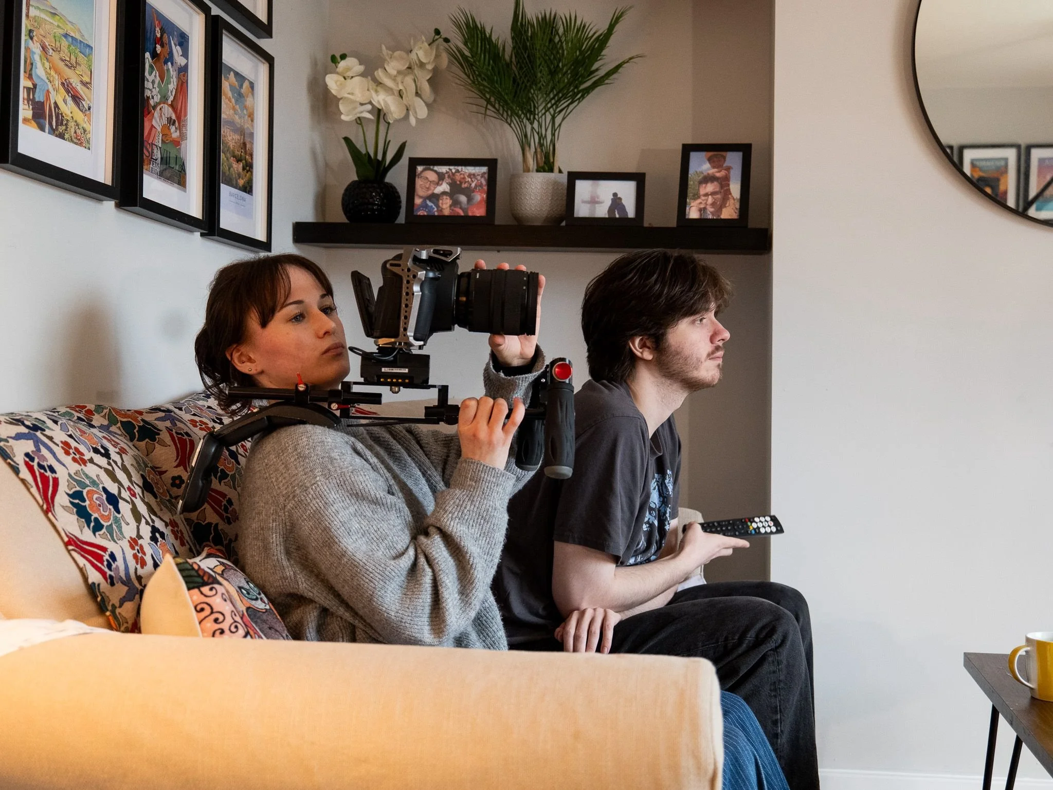 A woman filming with a camera while a man sits beside her on a couch, holding a remote control, in a living room with framed pictures and plants on the wall.