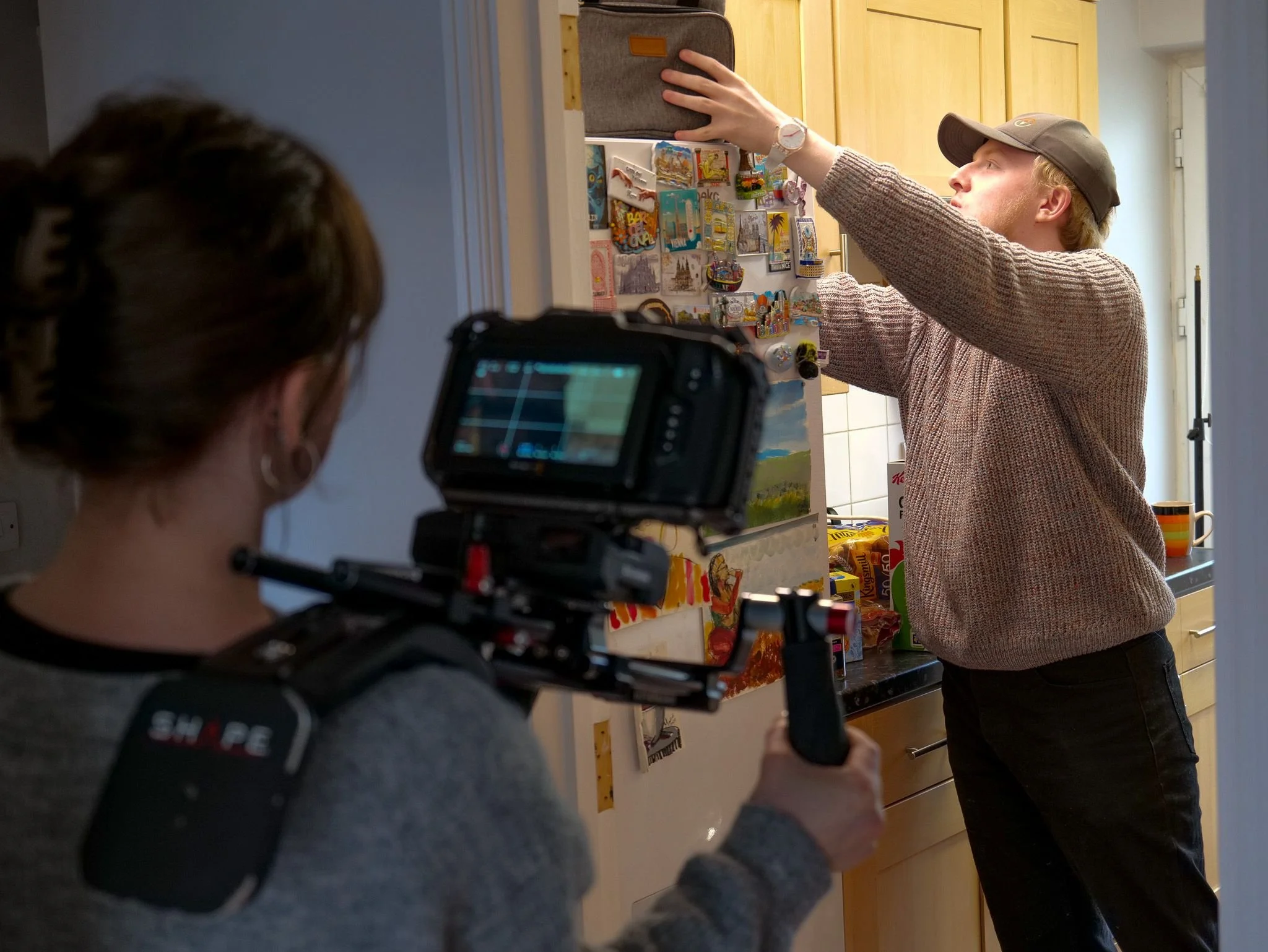 A woman is filming a man who is placing magnets on a refrigerator in a kitchen.