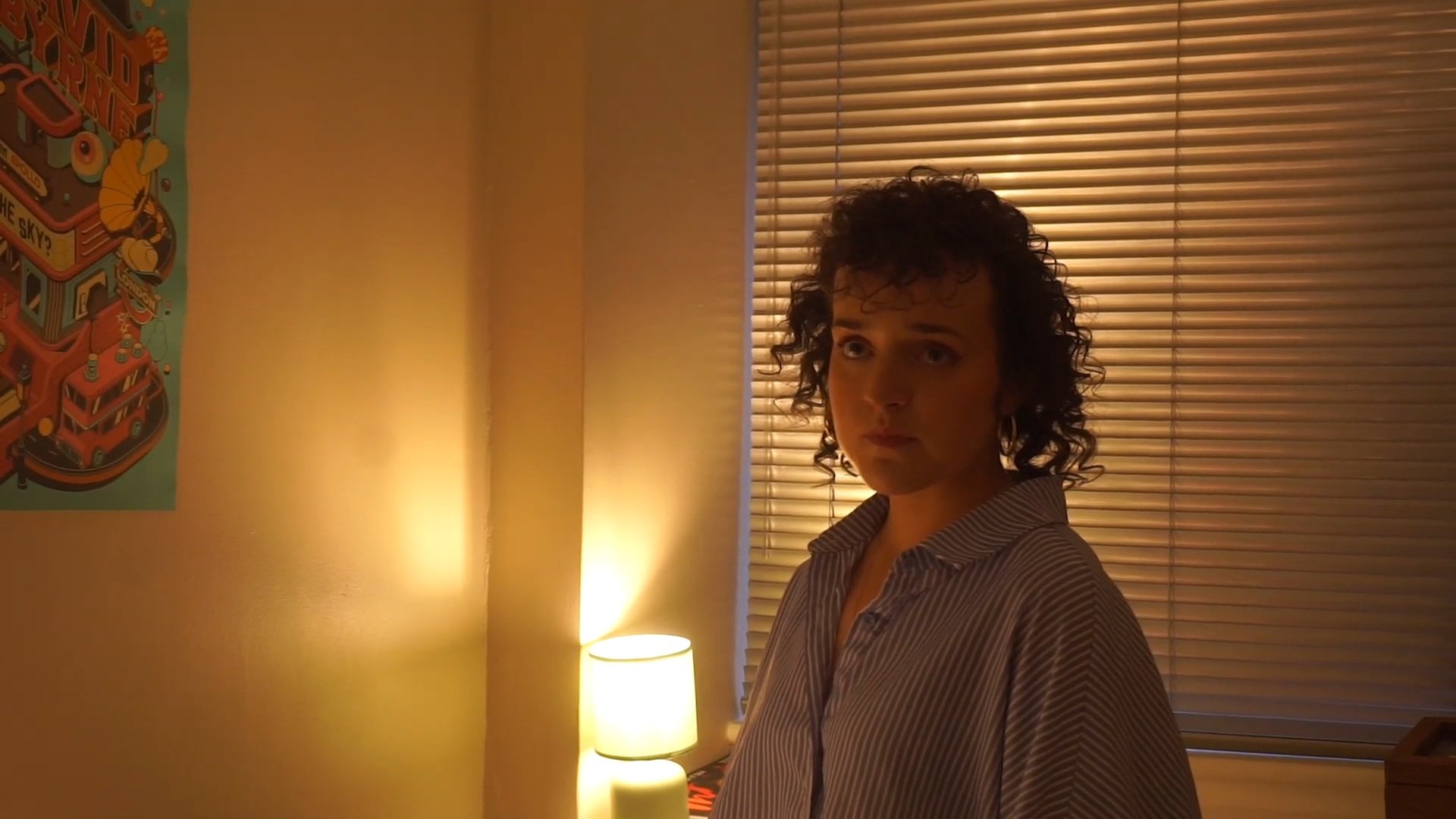 A woman with curly dark hair and striped shirt standing in a dimly lit room with a lamp and wooden blinds.