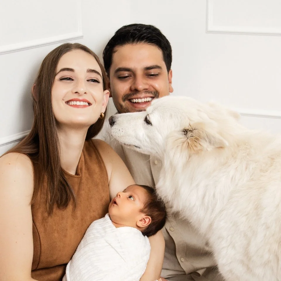 A happy family of three with their baby and large white dog, all smiling and snuggling together in a cozy indoor setting.