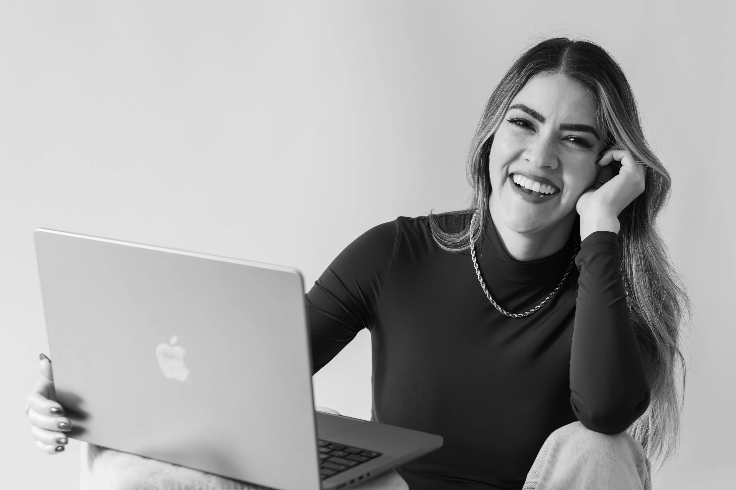 A young woman with long hair smiling and holding a laptop, sitting on the floor against a plain wall.