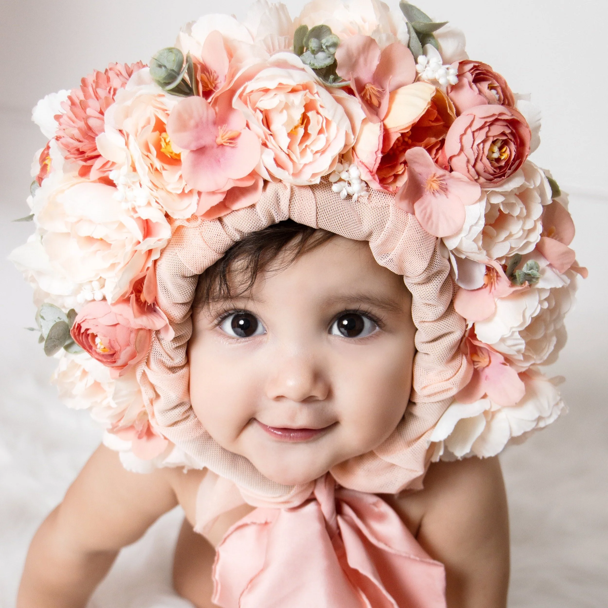 A smiling baby girl wearing a large floral bonnet with pink and white flowers.