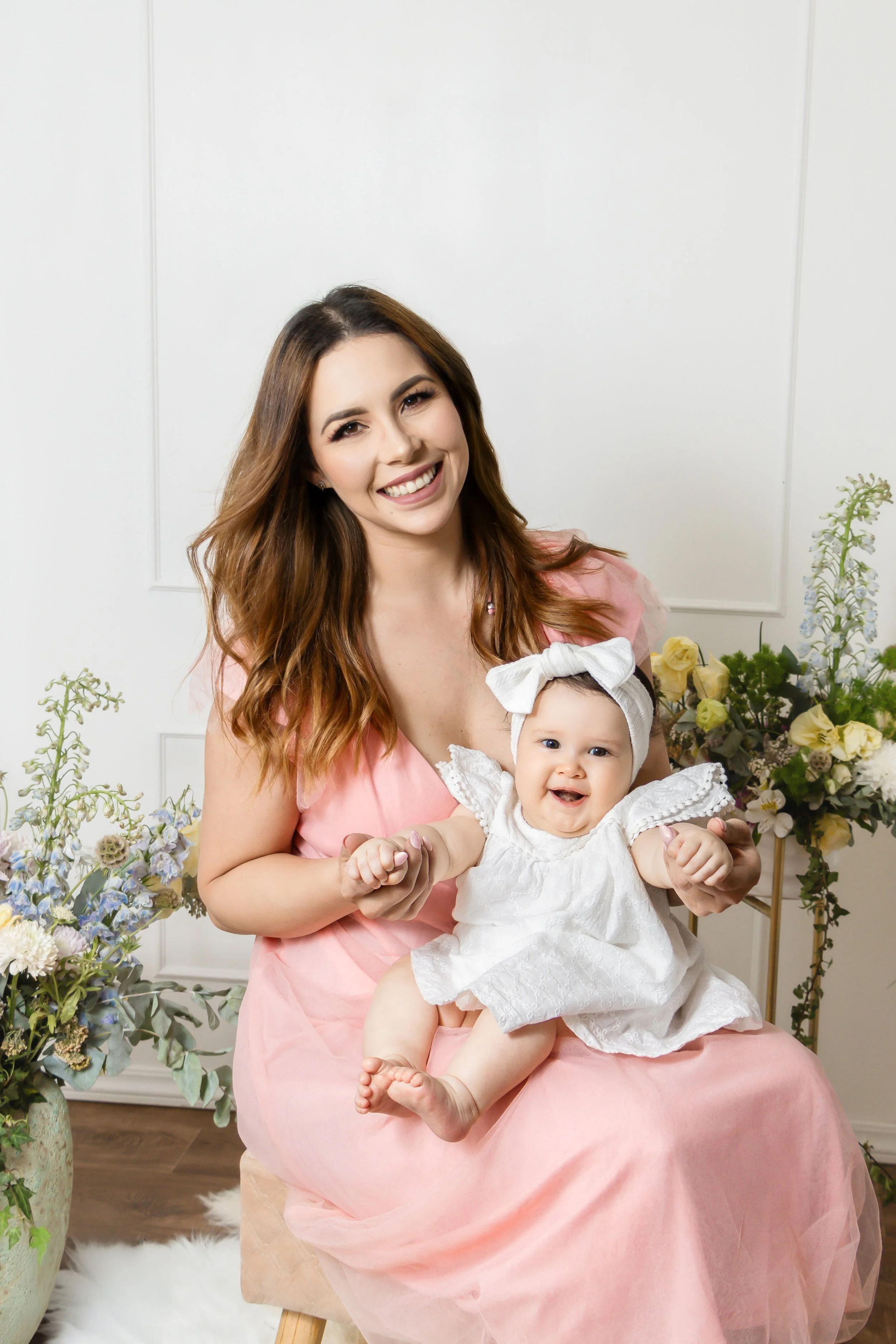 Smiling woman in pink dress holding a laughing baby girl in white dress and headband, seated in a floral decorated room.