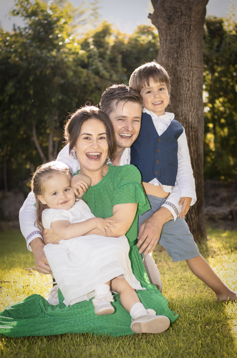Family of four smiling and laughing outdoors in a park near a tree during daytime, with sunlight filtering through the leaves.