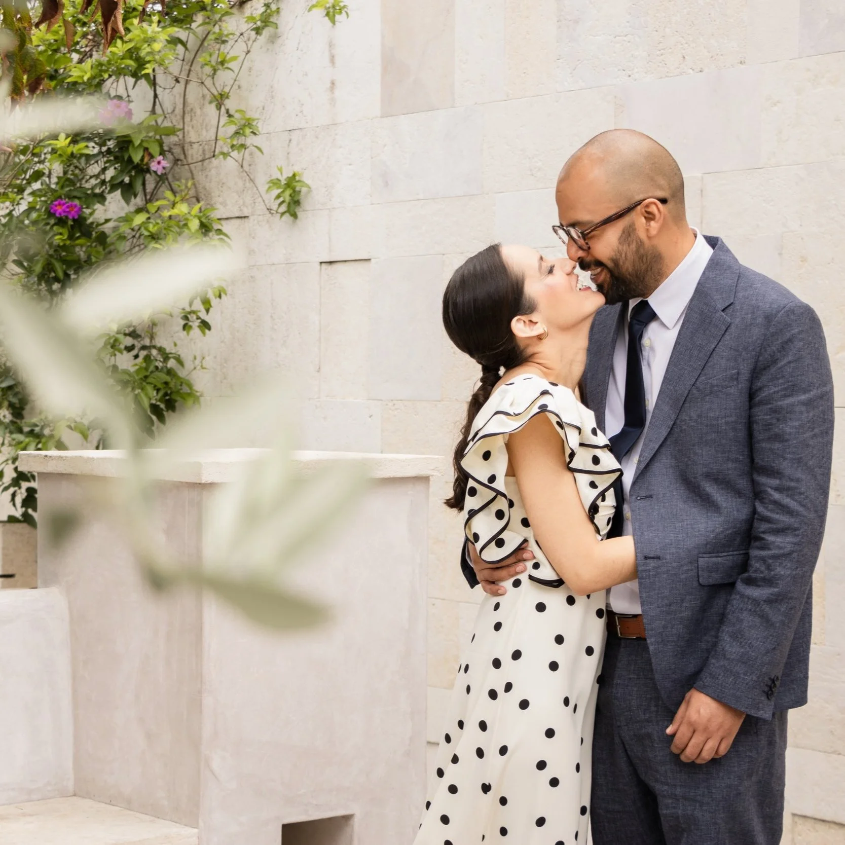 A couple wearing formal attire sharing a loving moment, standing close to each other against a light stone wall, with some greenery in the foreground.