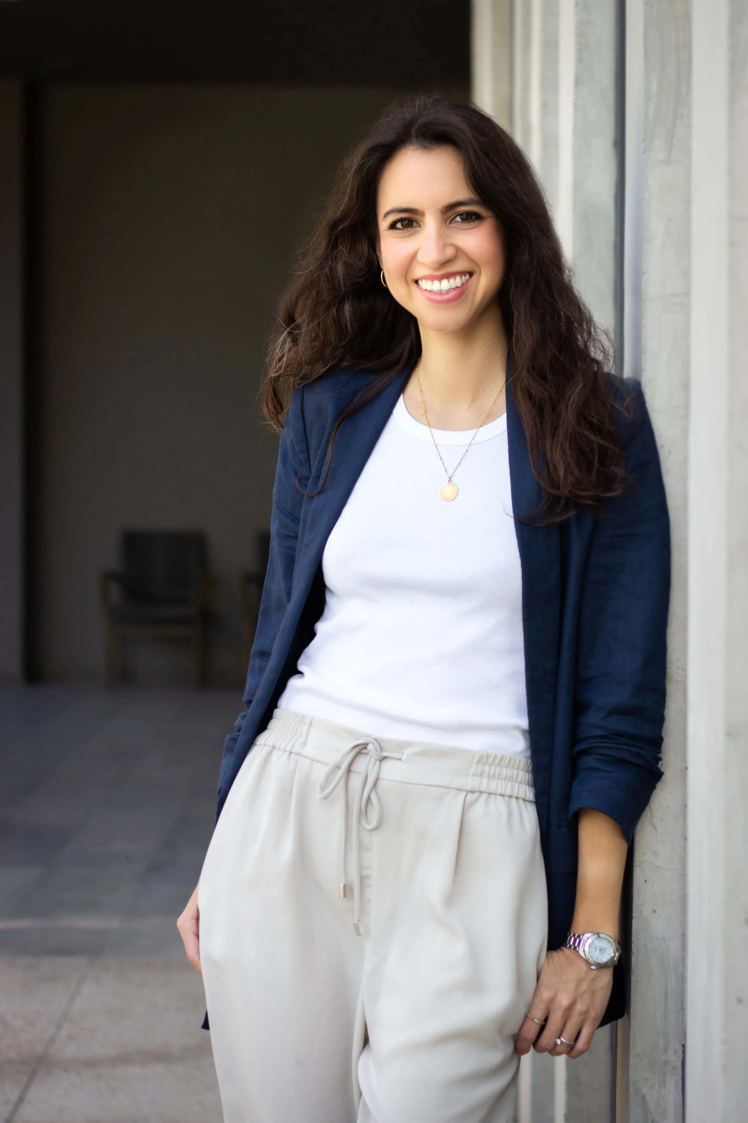 A woman with long brown hair, wearing a navy blazer, white t-shirt, beige drawstring pants, and a watch, standing outdoors and smiling.
