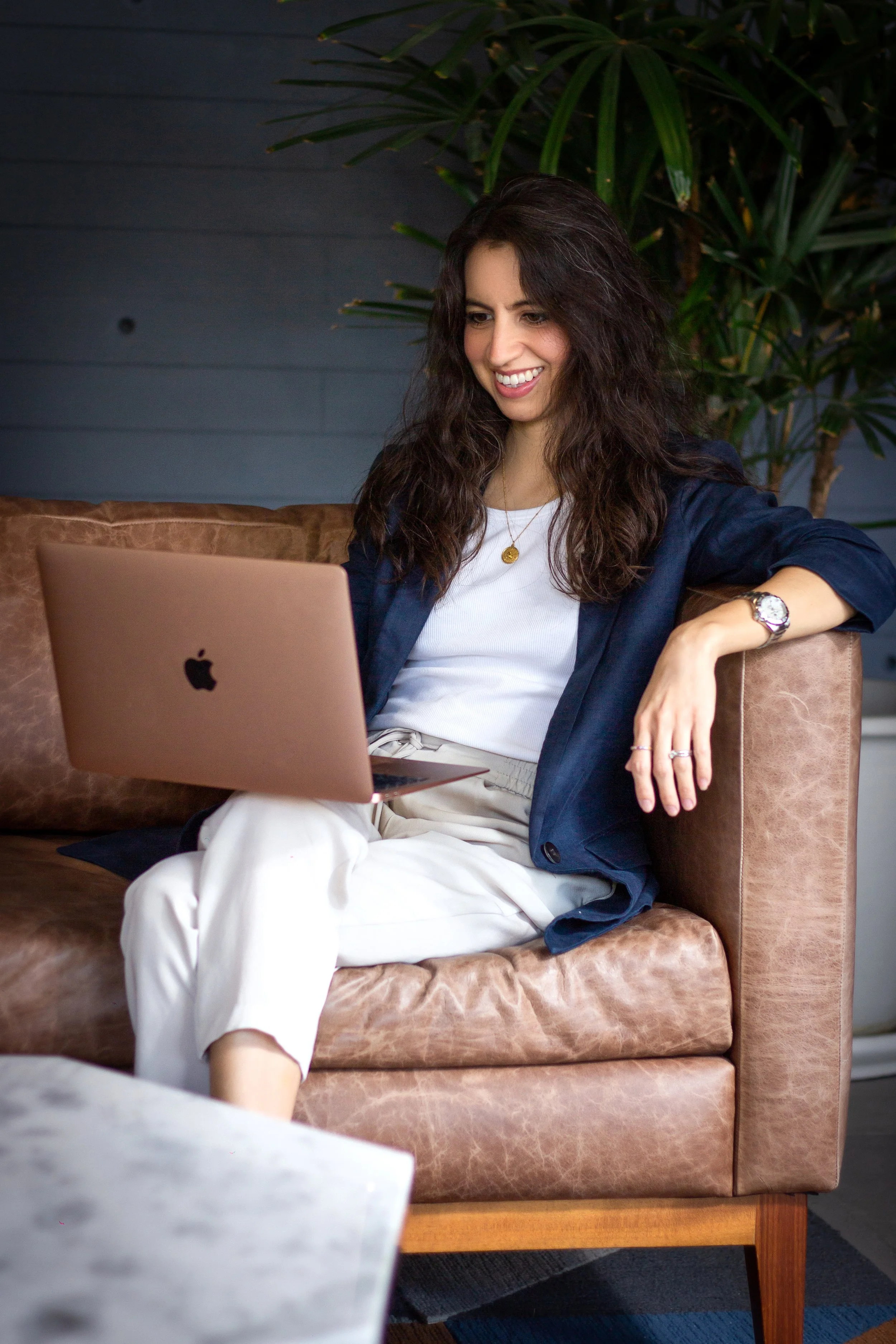 A woman with long curly dark hair, wearing a white shirt, navy blazer, and white pants, sitting on a brown leather couch, smiling while using a rose gold MacBook, with large green plant in the background.
