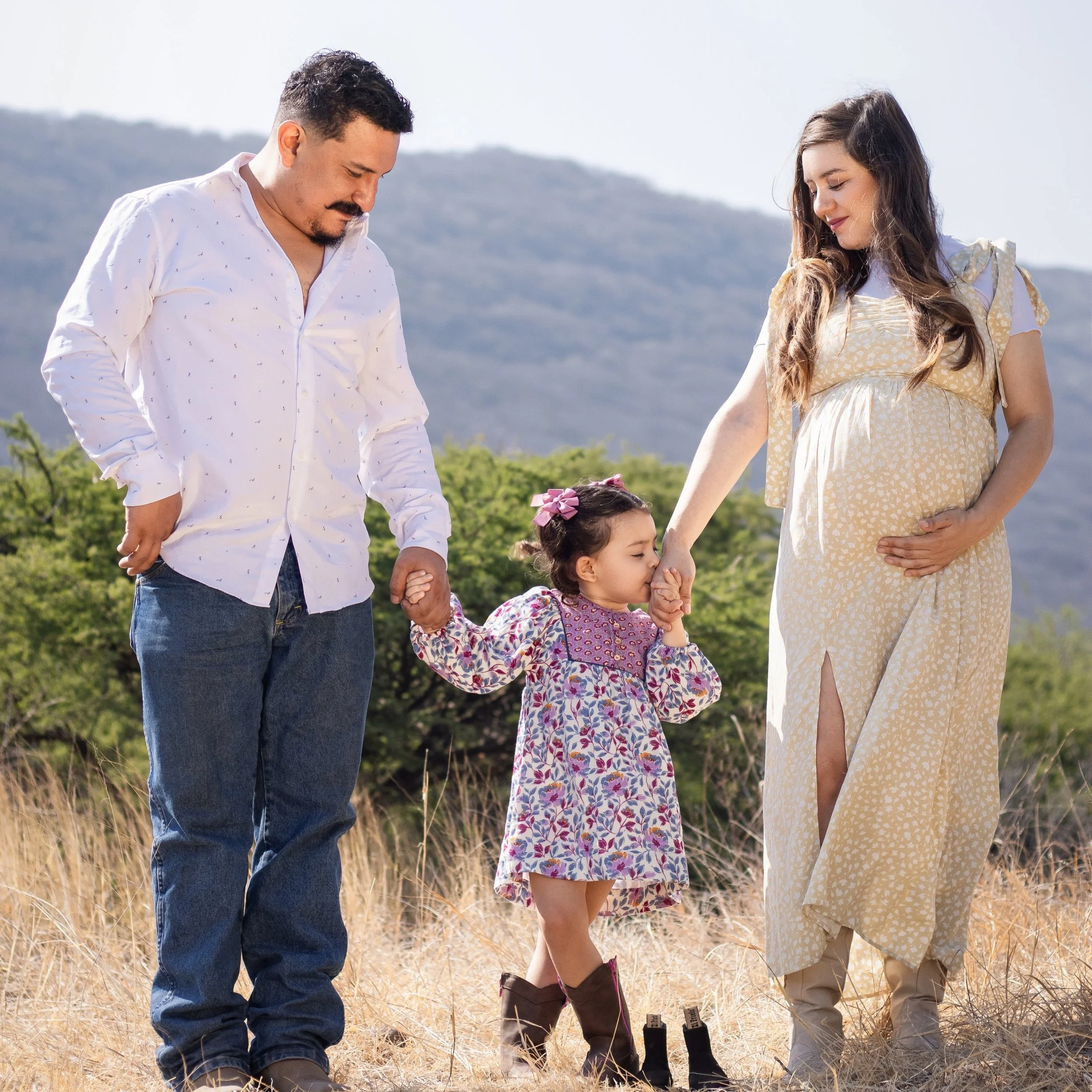 A pregnant woman and a man holding the hands of a young girl in an outdoor field with mountains in the background.