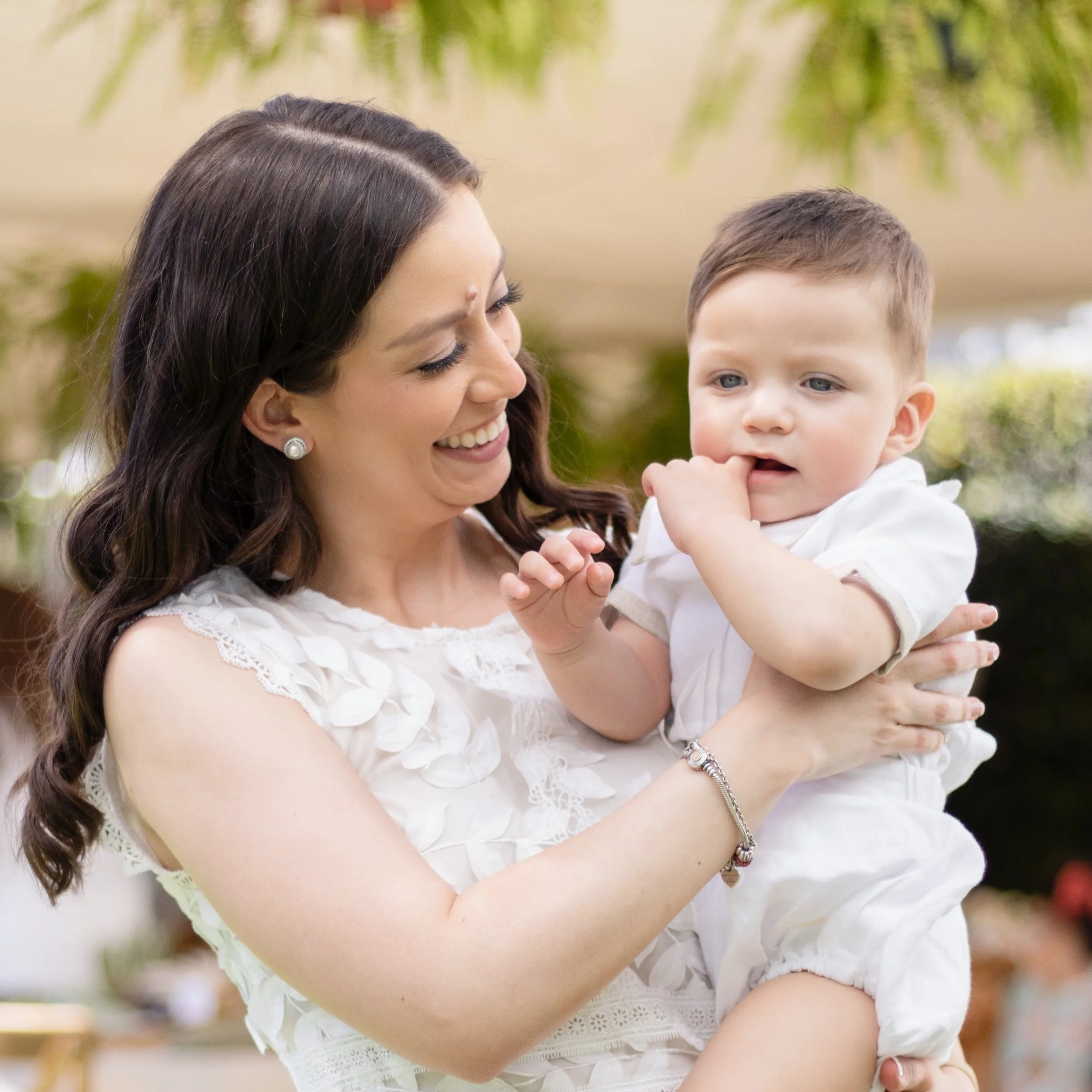 A woman with dark brown hair holding a young boy, both smiling and looking at each other, outdoors with blurred greenery in the background.