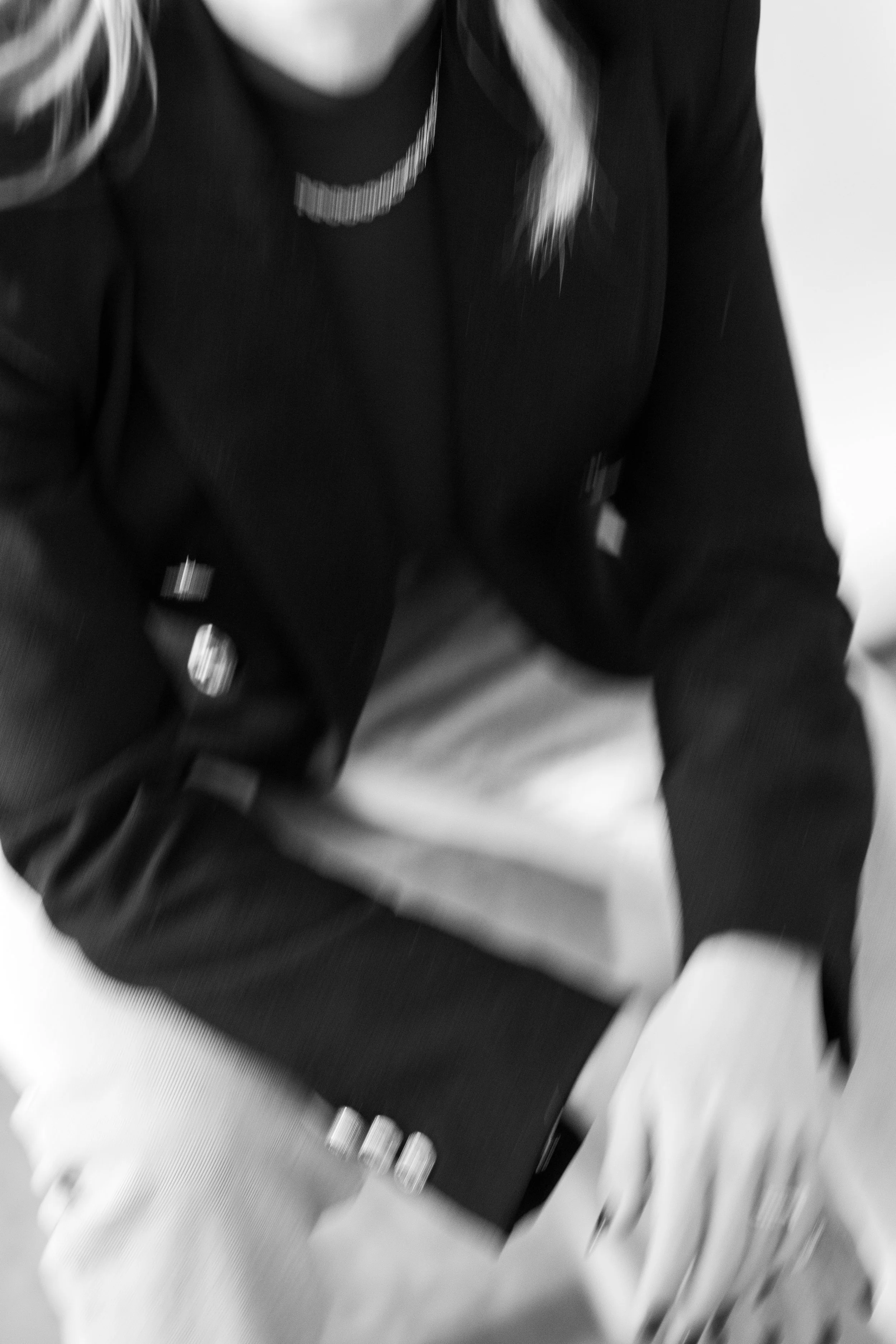 Black and white photo of a person wearing a dark blazer with a pocket square, light-colored pants, and a beaded necklace, sitting with their hands resting on their lap.
