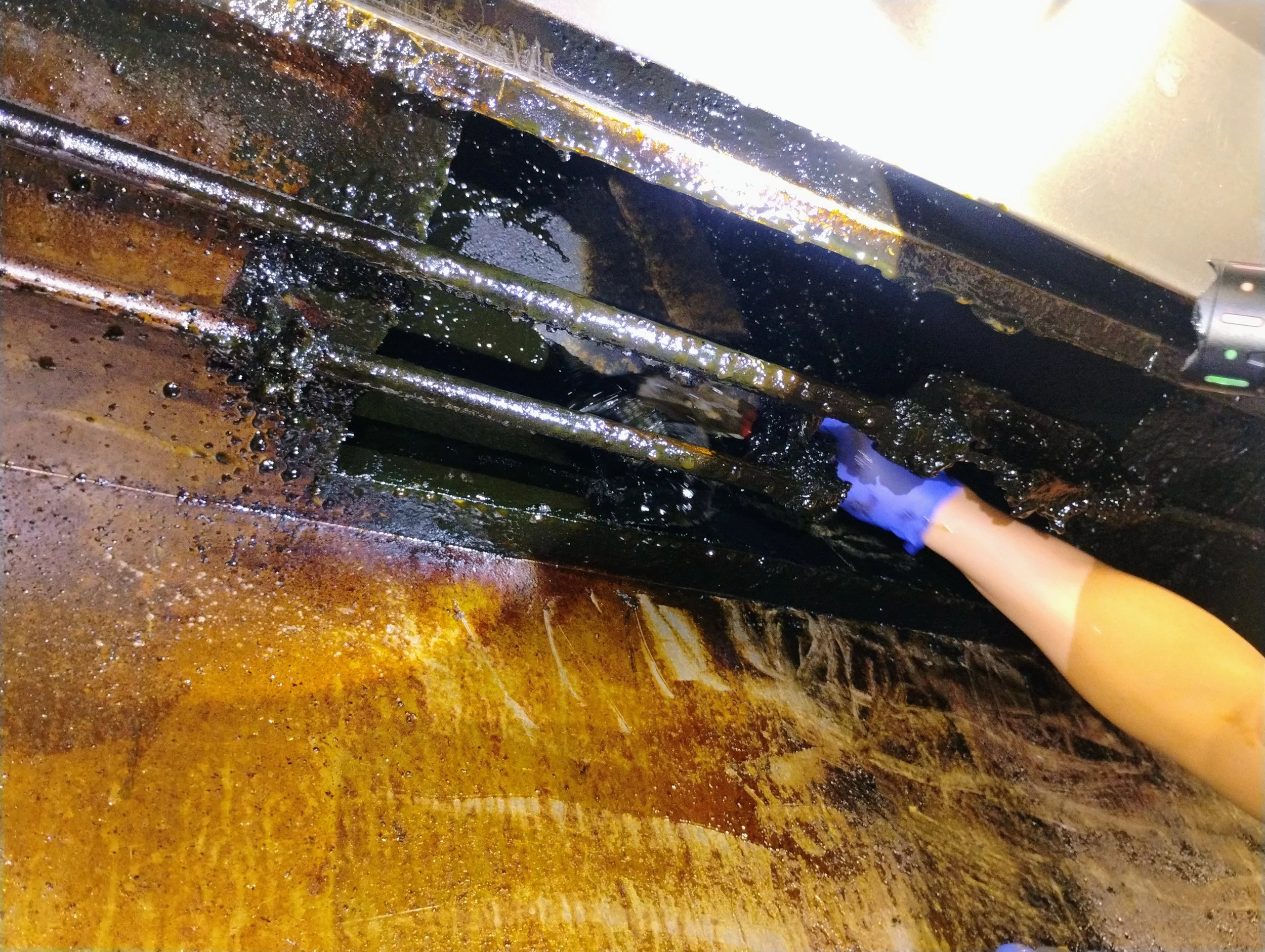 A person using a cleaning tool with a blue glove to scrub a greasy, blackened surface inside a kitchen stove or oven.