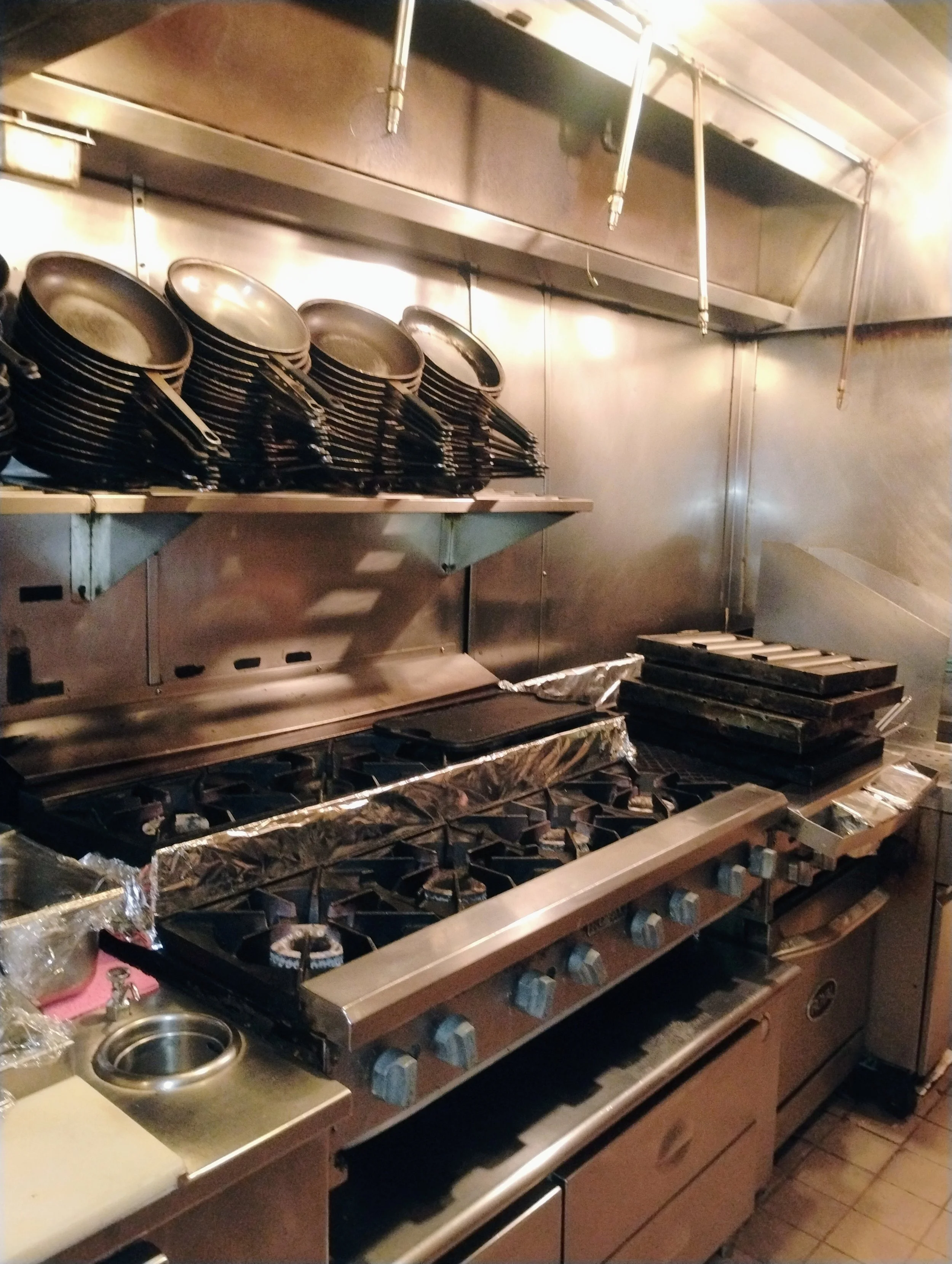 Commercial kitchen with stacked frying pans on a shelf, stacked grills or griddles, and a stovetop with burners, all made of stainless steel.