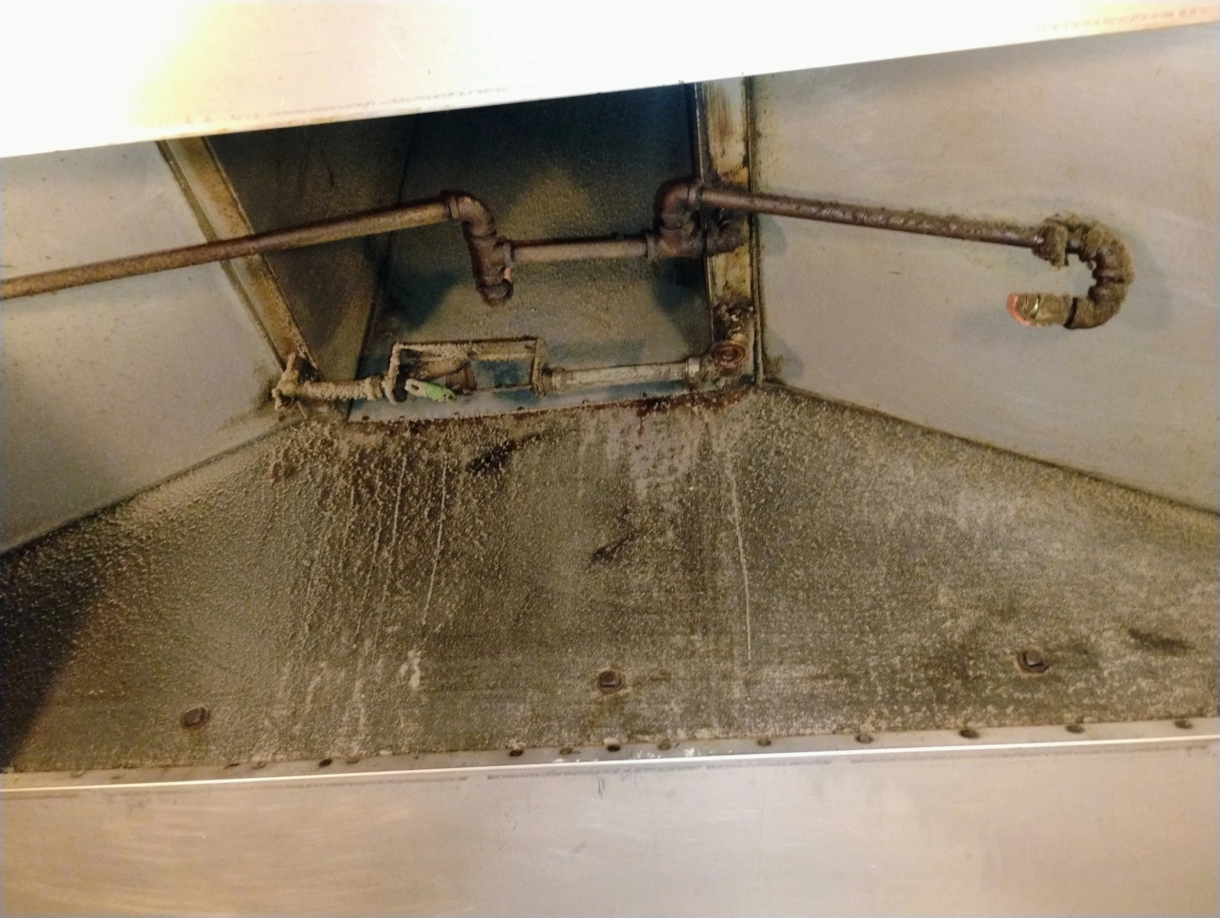 View of dirty storage compartment with rusted pipes and accumulated grime.