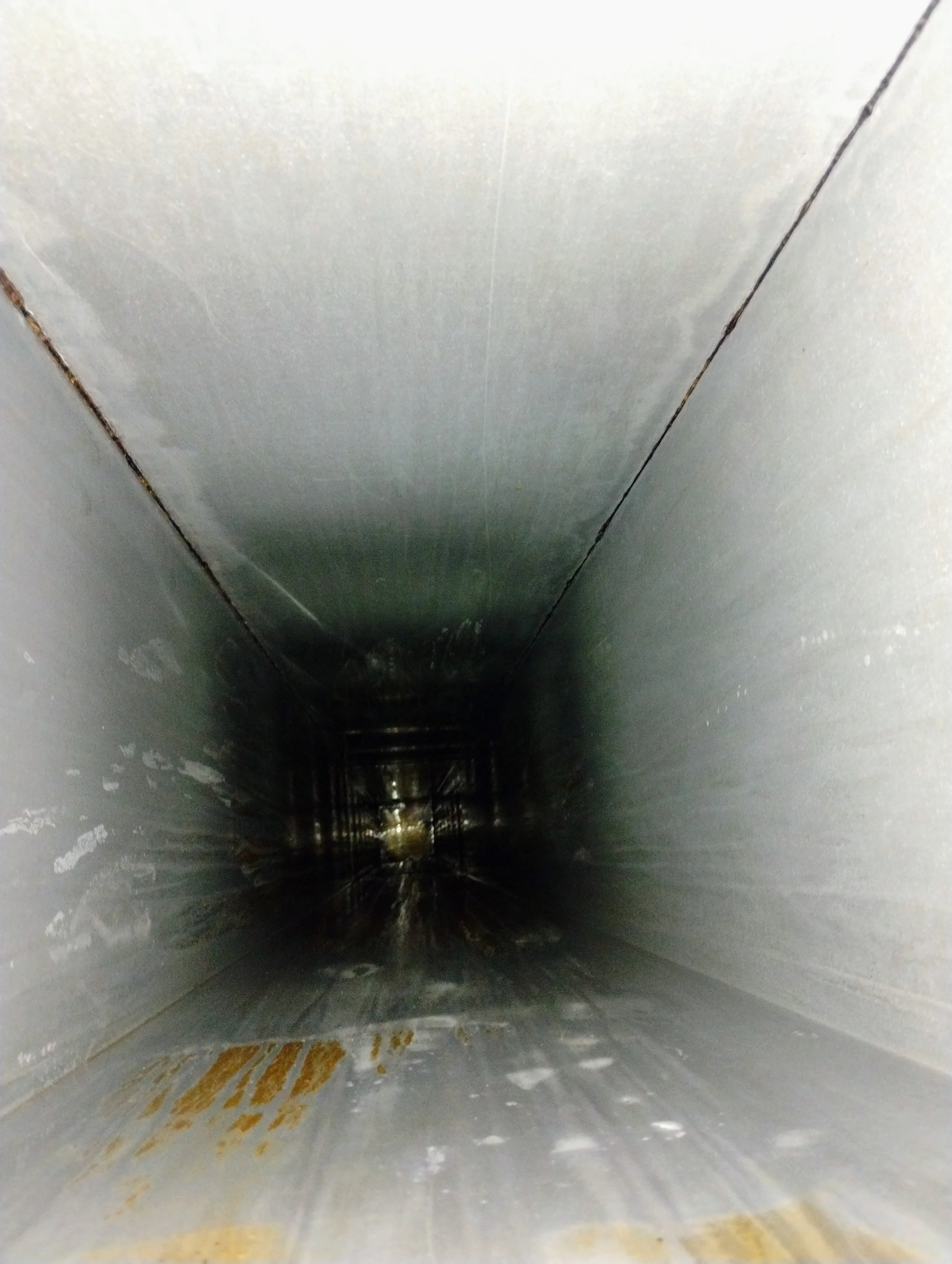 Inside a metallic pipe or duct, looking inward, with dark and shiny surfaces and some corrosion or rust at the edges.