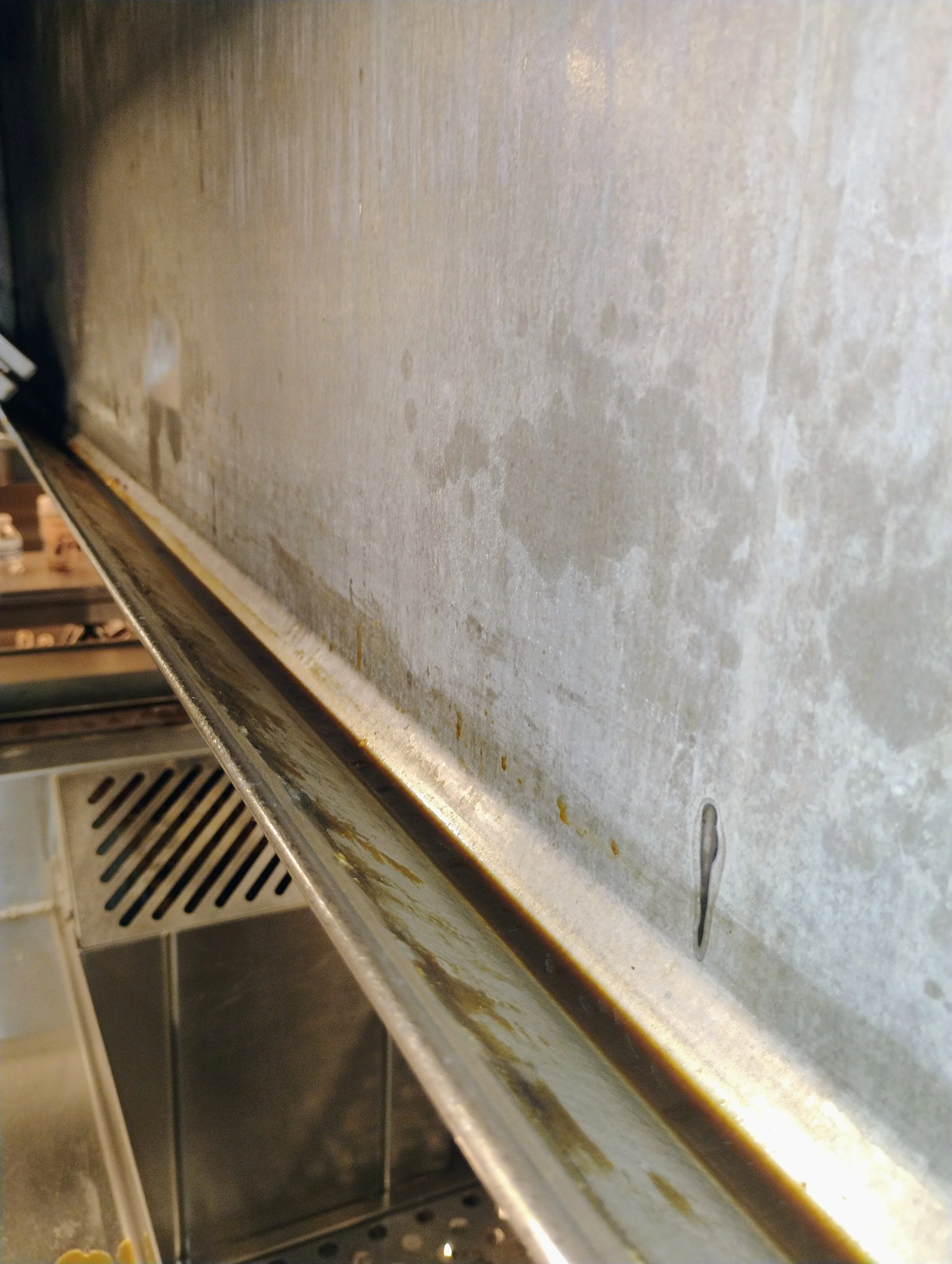 Close-up of a greasy, stained stainless steel surface in a kitchen, possibly a oven or appliance with food residue and rust.