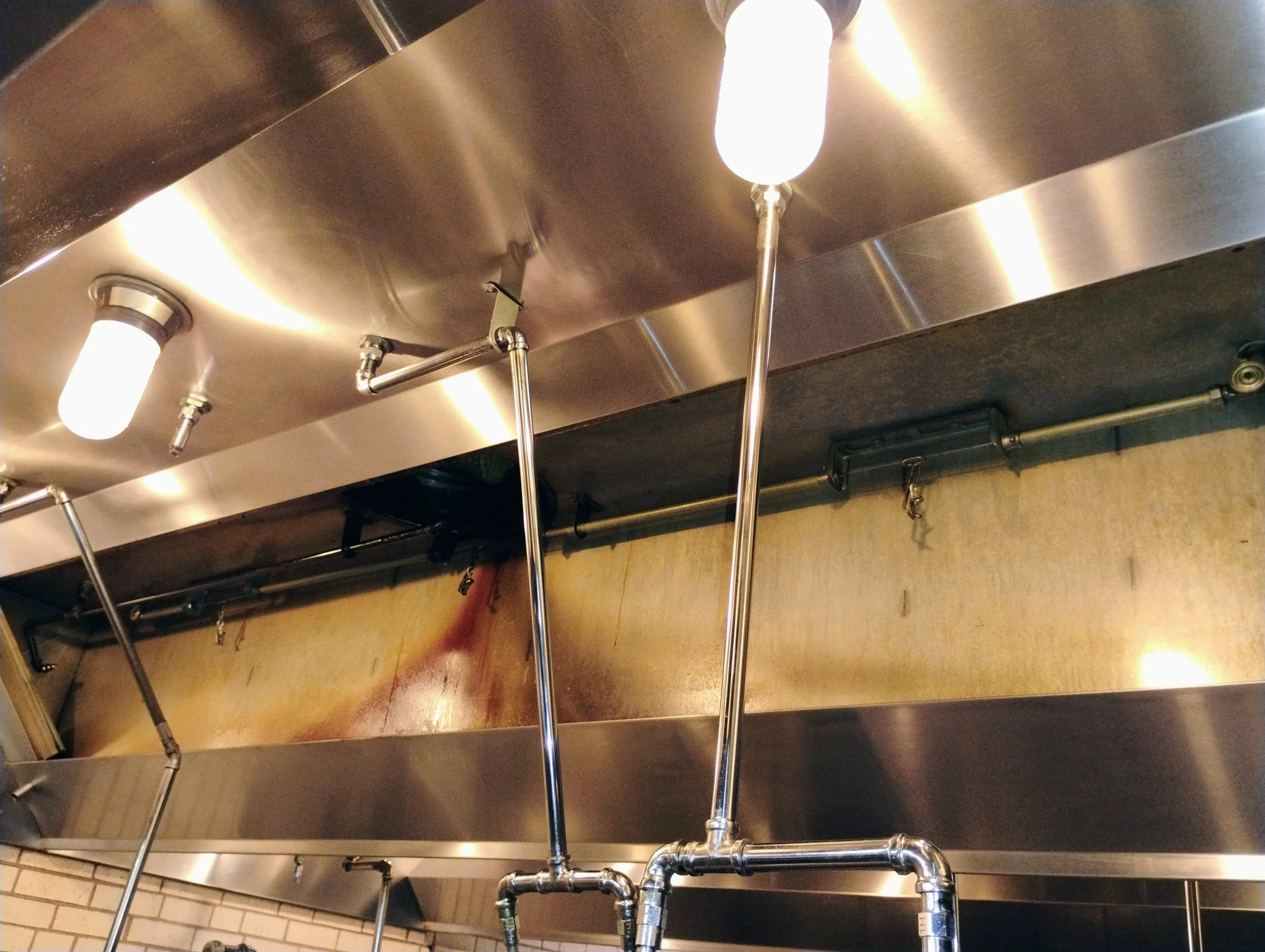 View of a commercial kitchen ceiling with stainless steel panels, fluorescent lights, copper pipes, and metal support structures.