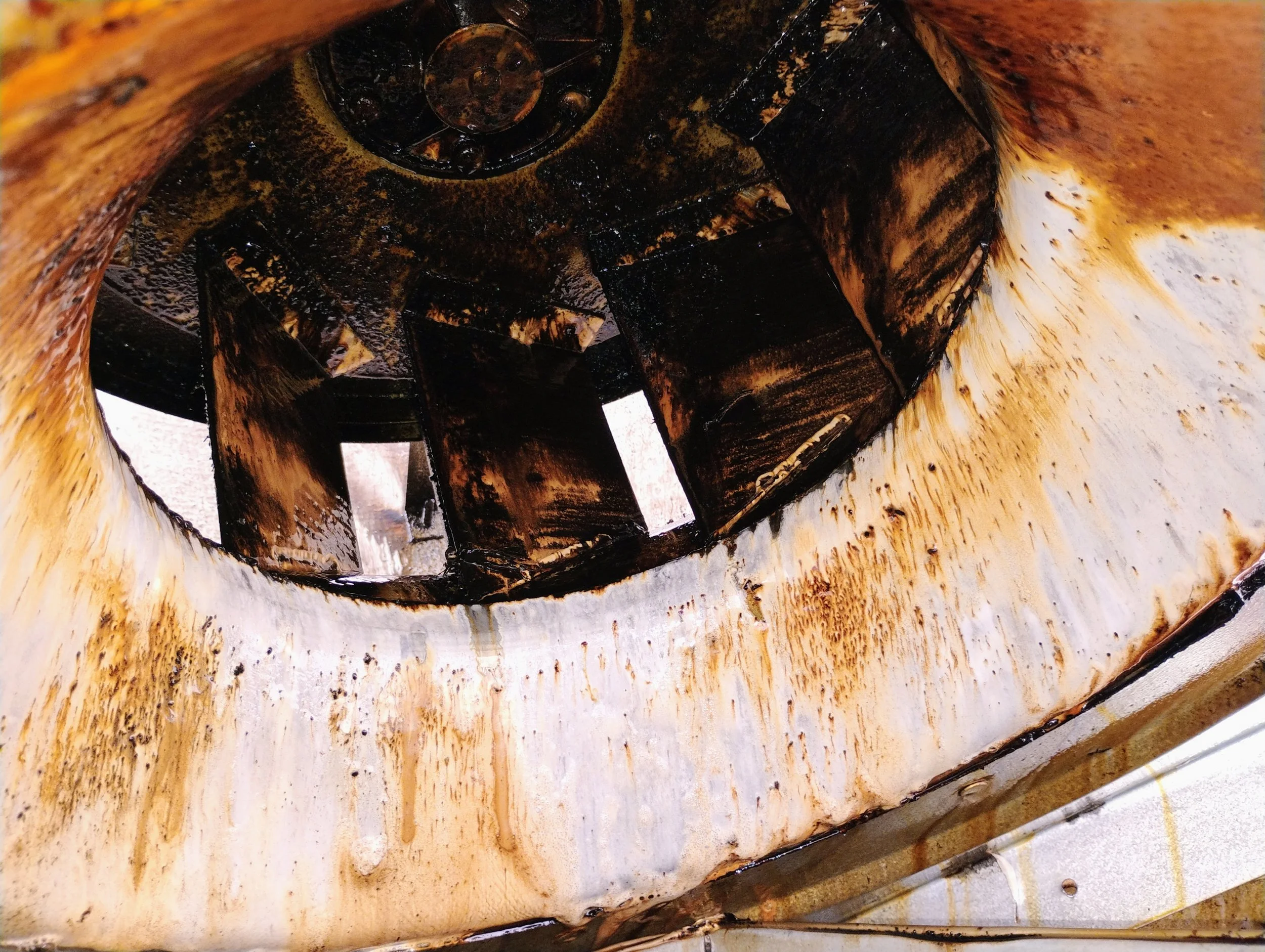 Close-up of a rusty, oily mechanical part, possibly a gear or wheel with a central hub and surrounding metal surfaces stained with oil or grease.