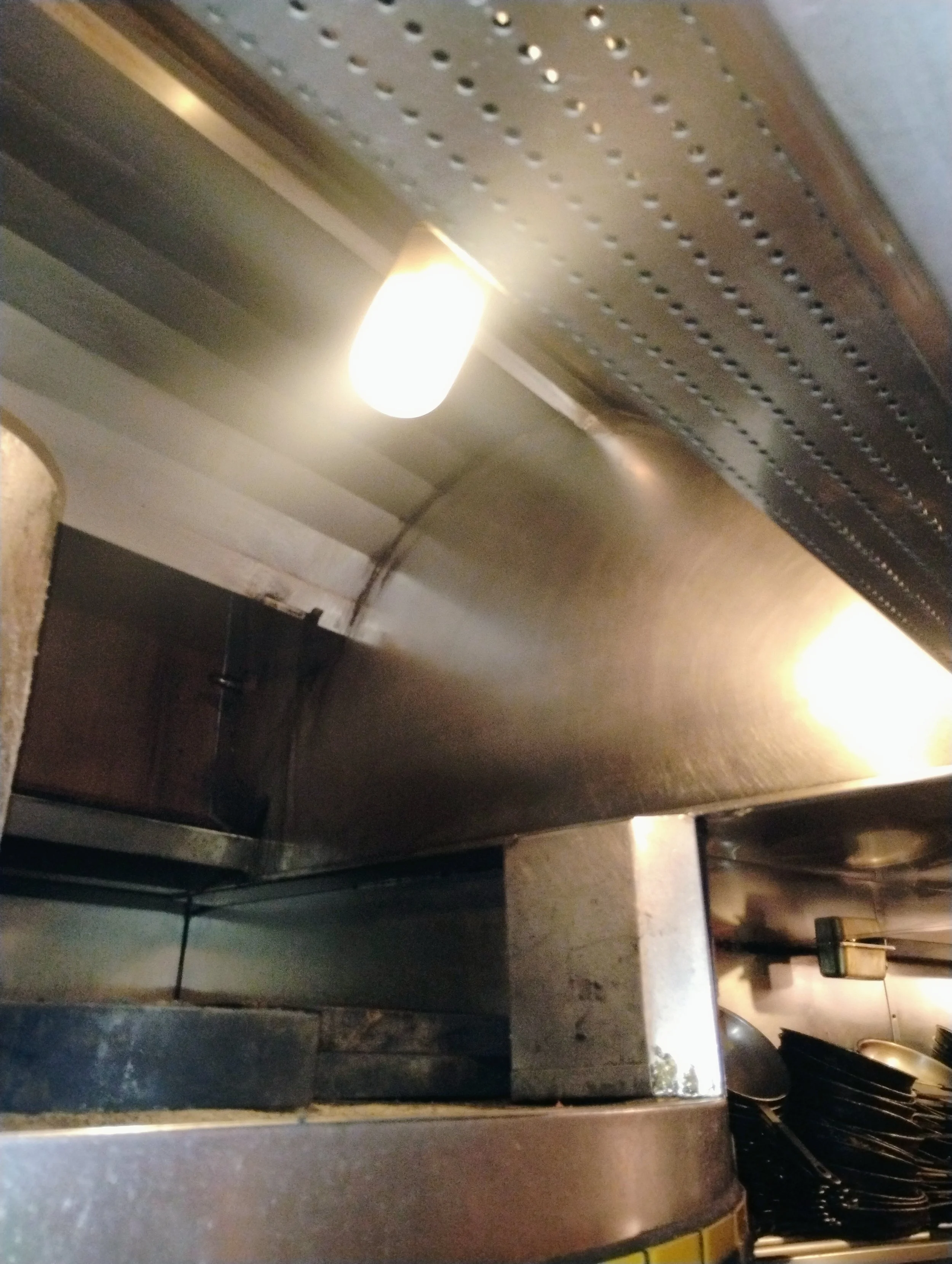 Close-up of a commercial kitchen stainless steel exhaust hood with lighting, showing the underside and some kitchenware in the background.