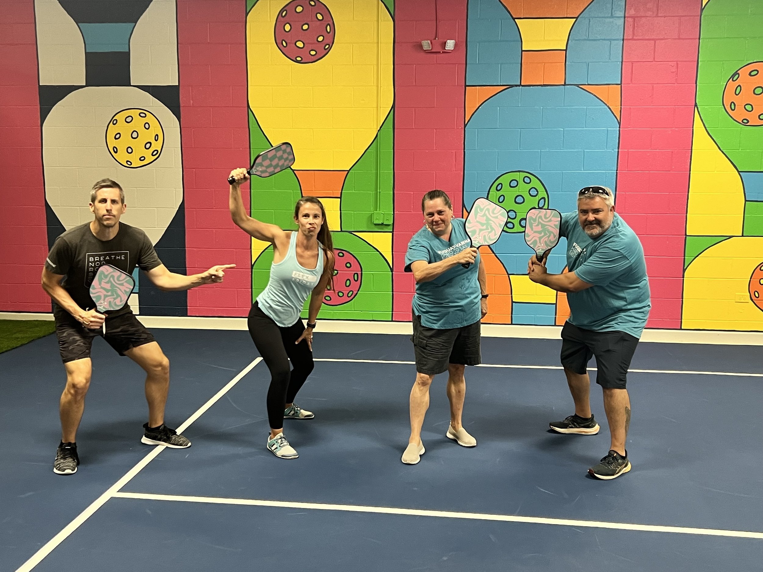 Four adults posing on a pickleball court, holding paddles, with a colorful, abstract mural background.