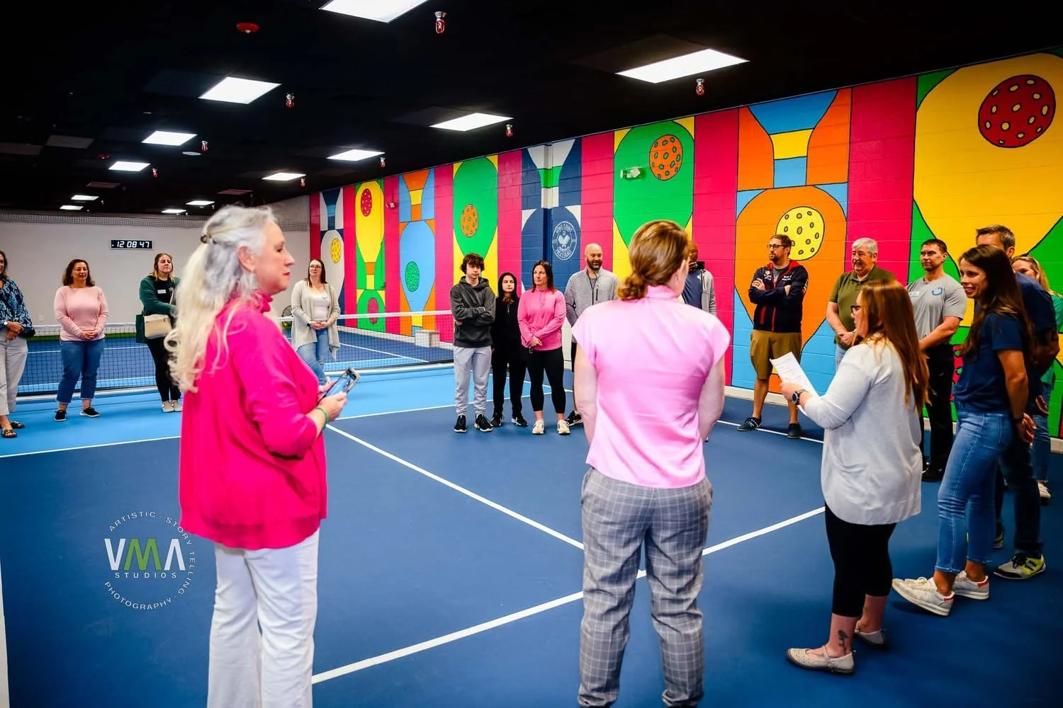 Group of people gathered in an indoor tennis court, listening to a woman speak. The court has a colorful, abstract mural on the wall and a digital clock displaying the time.