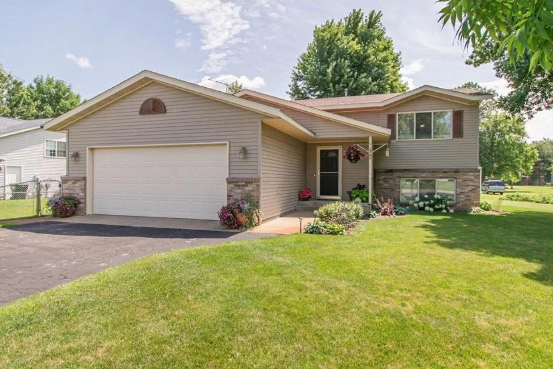 A two-story suburban house with beige siding and brick accents, a white garage door, and a manicured lawn with garden flowers and hanging baskets.