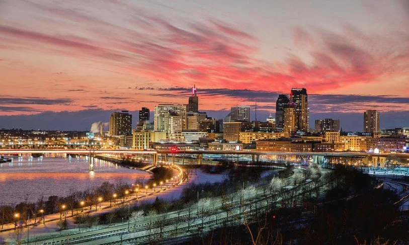 City skyline at sunset with a river, bridge, and illuminated buildings.