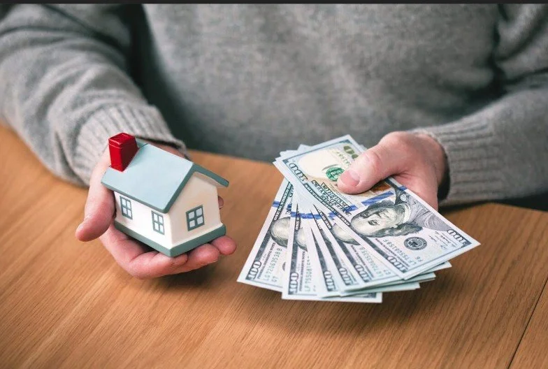 Person holding a small model house in one hand and several 100-dollar bills in the other.