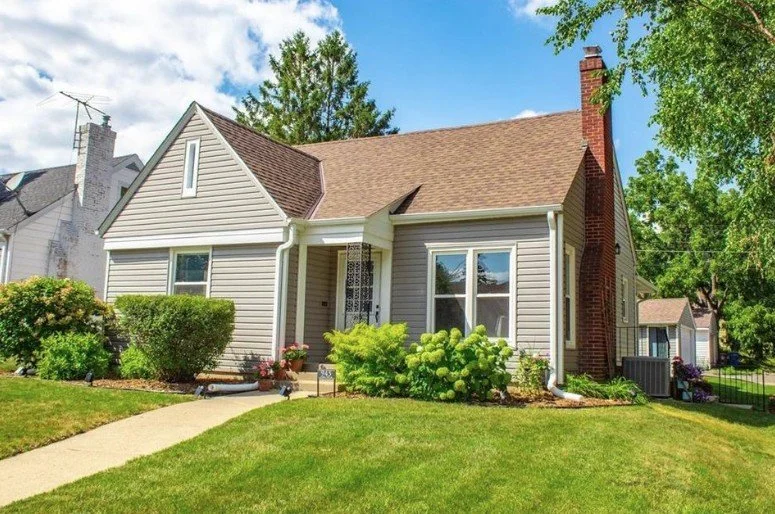 A small, charming house with beige siding, a brown shingle roof, and a red brick chimney. The front yard features a curved walkway, green lawn, and shrubs. There are trees in the background under a bright blue sky.