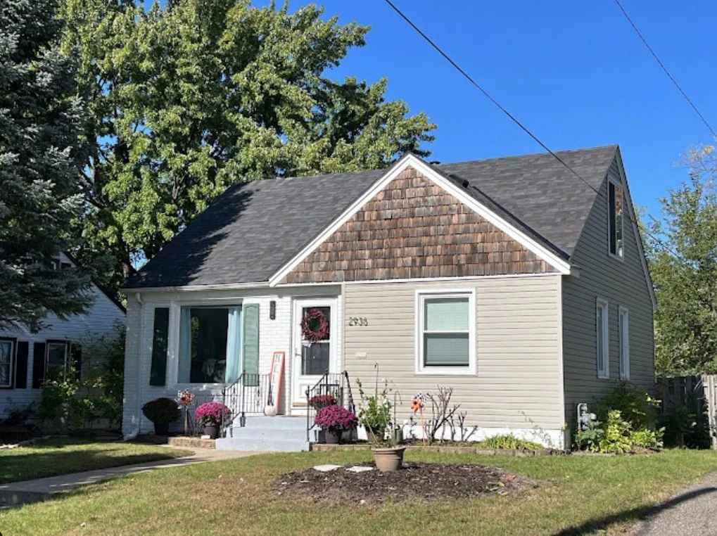 A house with beige siding and a gabled roof, decorated with potted plants and flowers in front, under a large tree on a clear day.