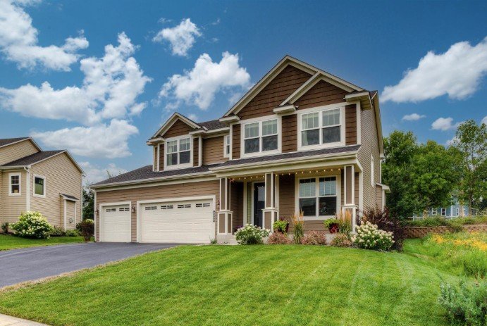 A two-story suburban house with a brown exterior, white trim, and a three-car garage, surrounded by a green lawn and landscaped garden with bushes and flowers.