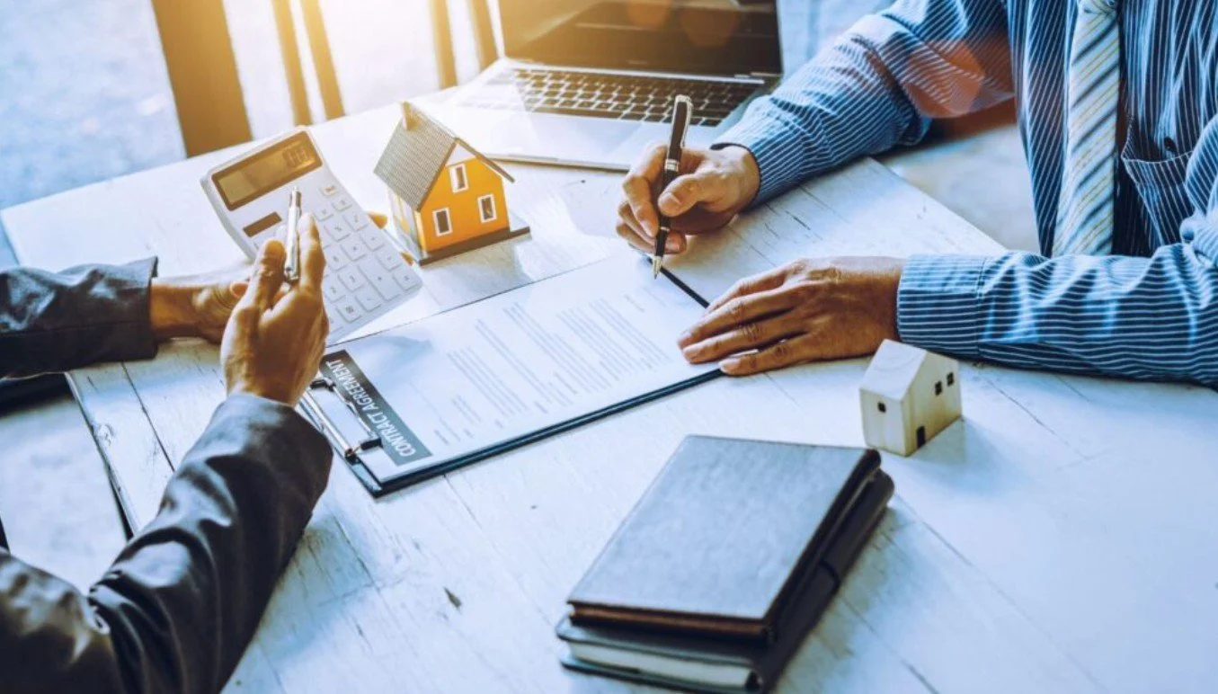 Two people sit at a table discussing real estate or home buying, with documents, small house models, a laptop, and a calculator on the table.