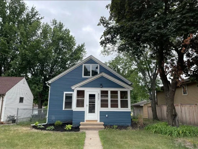 A blue, two-story house with a small front porch, surrounded by trees and neighboring houses.