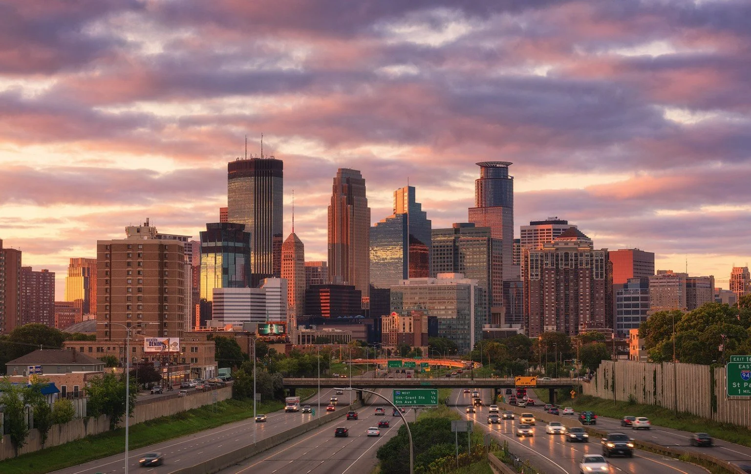City skyline at sunset with tall skyscrapers and traffic on a highway in the foreground.