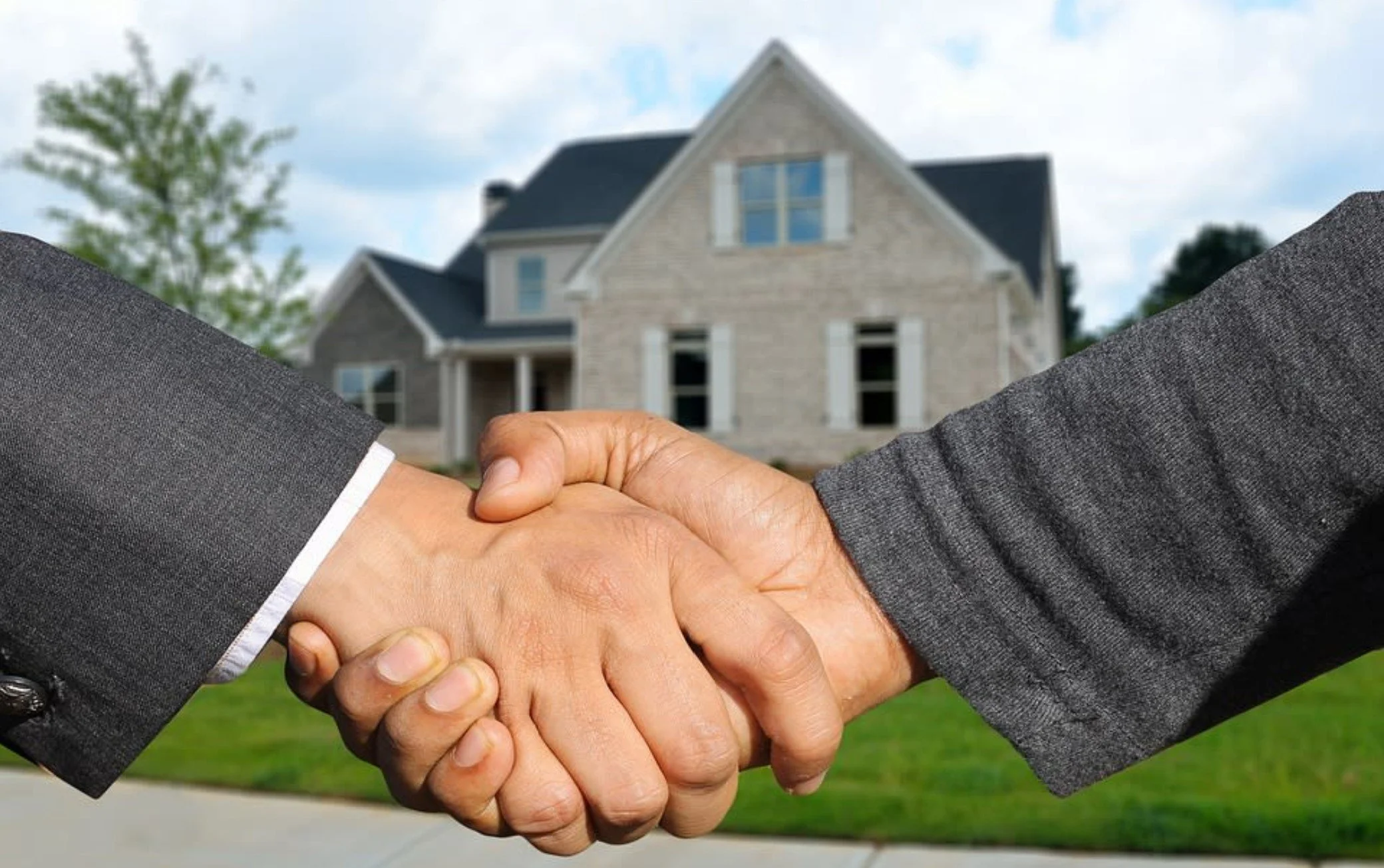A handshake between two people in business suits in front of a house, symbolizing a real estate agreement or closing deal.