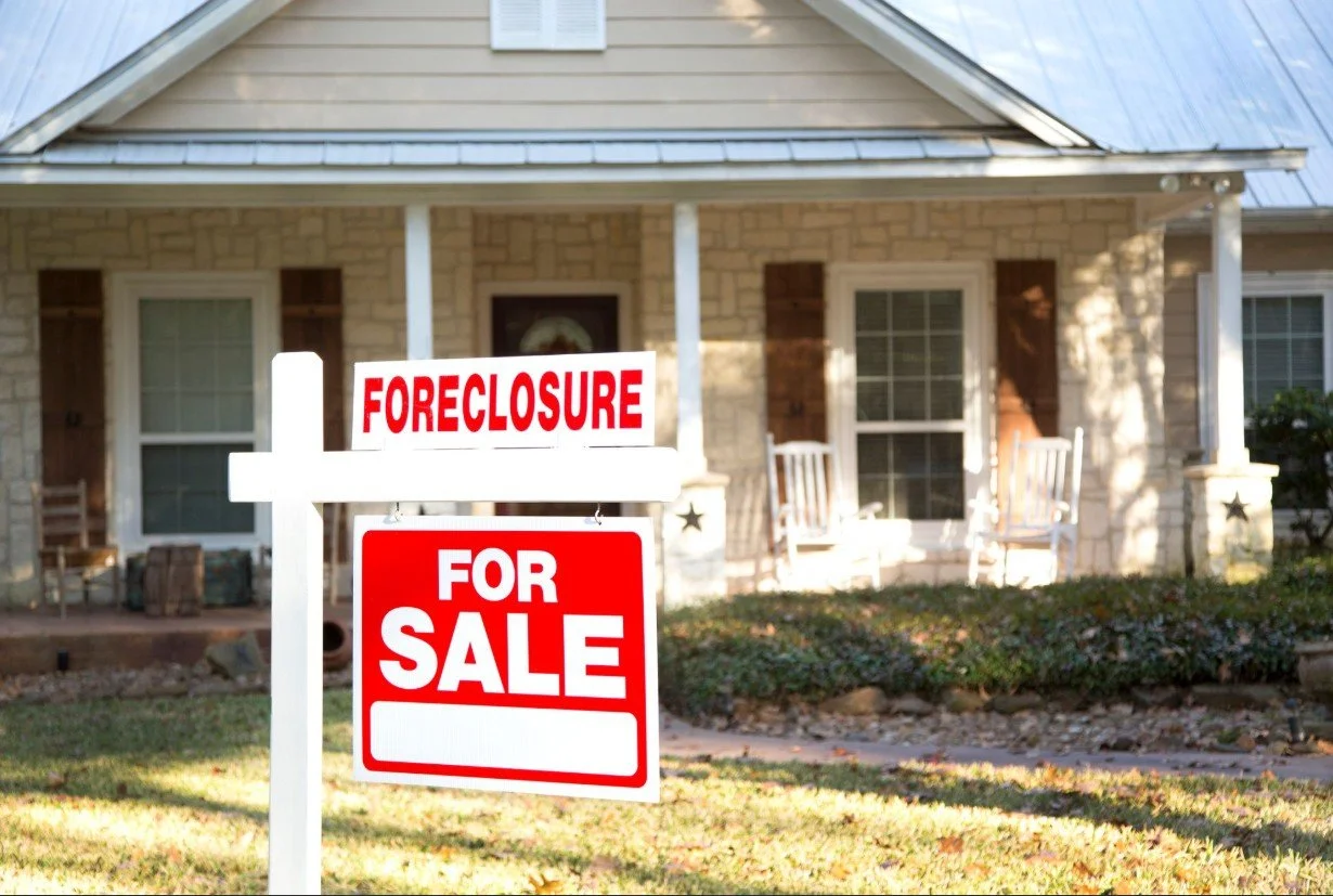 A residential house with a "Foreclosure" sign and a "For Sale" sign in the front yard.