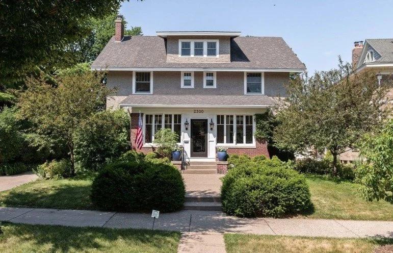 A large, gray, three-story house with a covered front porch and multiple windows, surrounded by greenery and shrubs, with a concrete sidewalk in front.