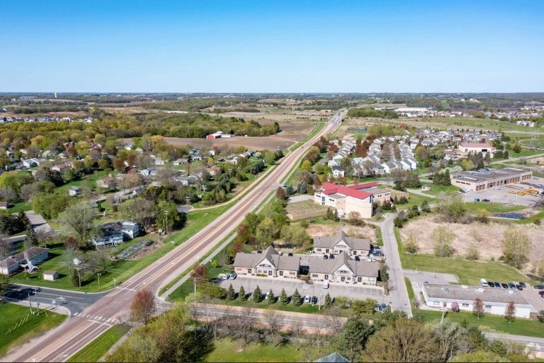 Aerial view of a suburban area with a highway passing through, surrounded by houses, trees, and open fields, under a clear blue sky.