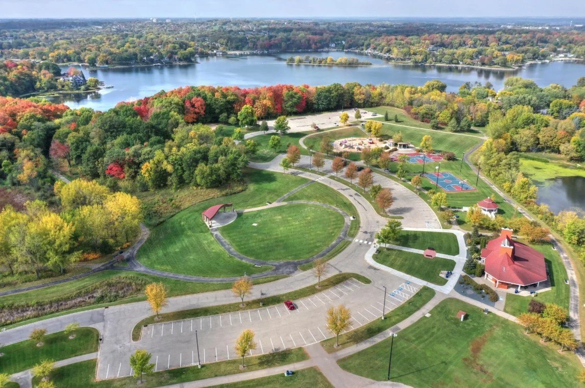 Aerial view of a park with green lawns, trees with autumn foliage, a lake in the background, and recreational facilities including tennis courts, playgrounds, and a building with a red roof.